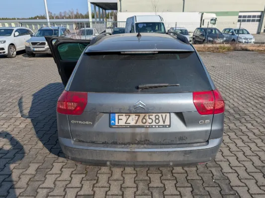 Z tyłu widoczny szary samochód Citroën C5 z otwartymi drzwiami bagażnika. Na tablicy rejestracyjnej widnieje numer FZ 7658V z literą A po lewej stronie. Samochód stoi na brukowanej powierzchni, w tle znajdują się inne pojazdy, budynek przemysłowy i ogrodzenie. Na tylnym szybie widoczny jest wycieraczka, a na bagażniku logo marki i oznaczenie C5.