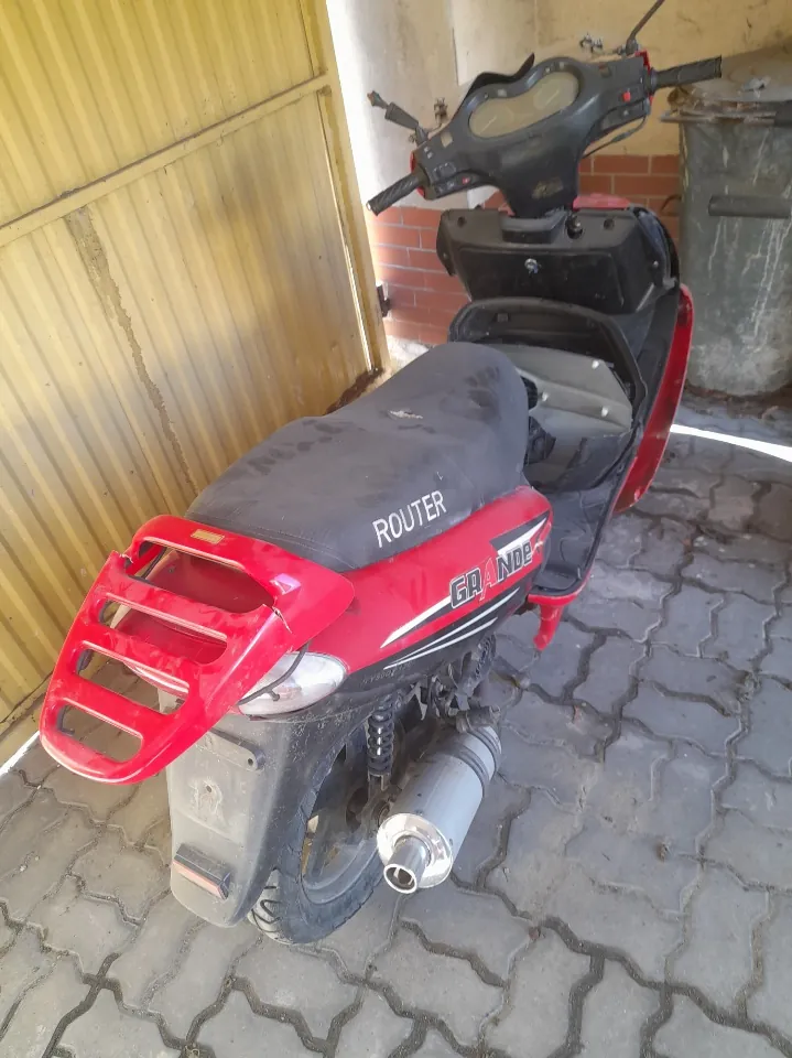 Czerwony skuter zaparkowany na brukowanej powierzchni. Skuter ma czarne siedzenie z białym napisem ROUTER oraz napisem GRANDE na czerwonej części kadłuba. W tle widoczna żółta metalowa brama, ceglana ściana i duży szary pojemnik. Skuter jest zwrócony tyłem do obserwatora, widoczne są tylna część, reflektor, wydech oraz kierownica.