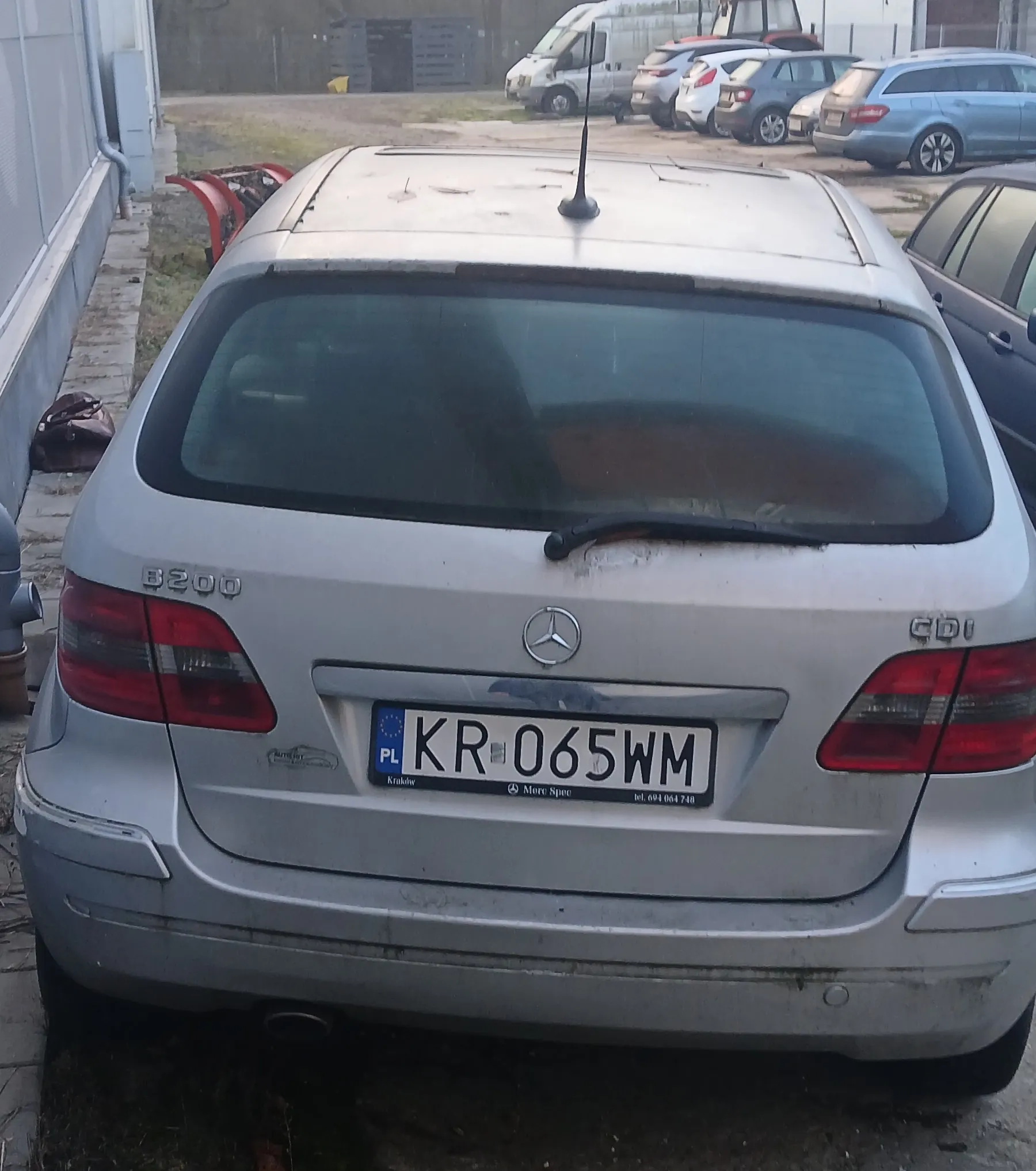 Tył srebrnego samochodu Mercedes-Benz z tablicą rejestracyjną KR-065WM (PL, Kraków). Na bagażniku widoczne oznaczenia 8200 i CDI, logo Mercedes-Benz oraz antena. Samochód zaparkowany na otwartym placu, w tle inne pojazdy, budynek po lewej stronie i trawa. Tablica rejestracyjna zawiera dodatkowo napis More Spec oraz numer telefonu 694 064 748.