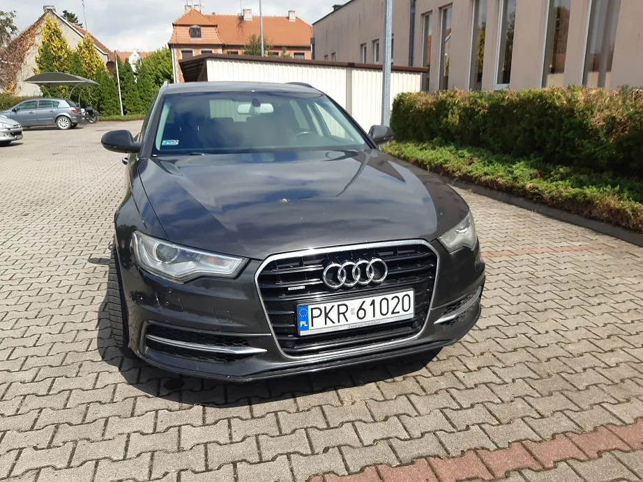 Czarny samochód marki Audi zaparkowany przodem do obiektywu na kostce brukowej. Na grillu widoczne są cztery pierścienie logo Audi oraz polska tablica rejestracyjna o numerze PKR 61020. W tle widać niskie budynki, zielone krzewy oraz inny zaparkowany samochód.