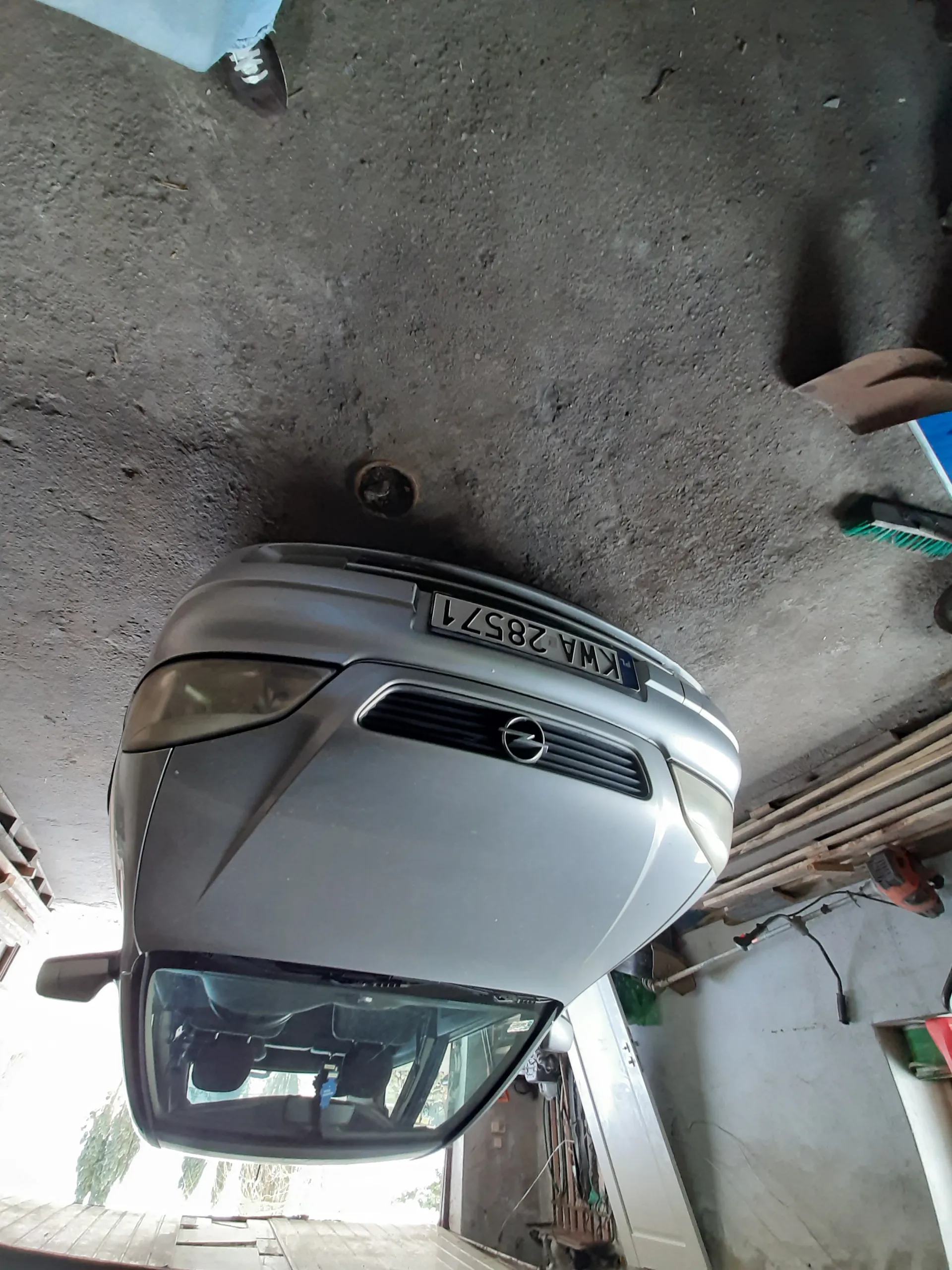 Srebrny samochód marki Opel z tablicą rejestracyjną KWA 28571 zaparkowany w garażu. Na betonowej podłodze widoczne narzędzia, deski oraz część nogi osoby w niebieskich dżinsach i czarnym butie. W tle otwarte drzwi garażu z widokiem na zieloną roślinność.