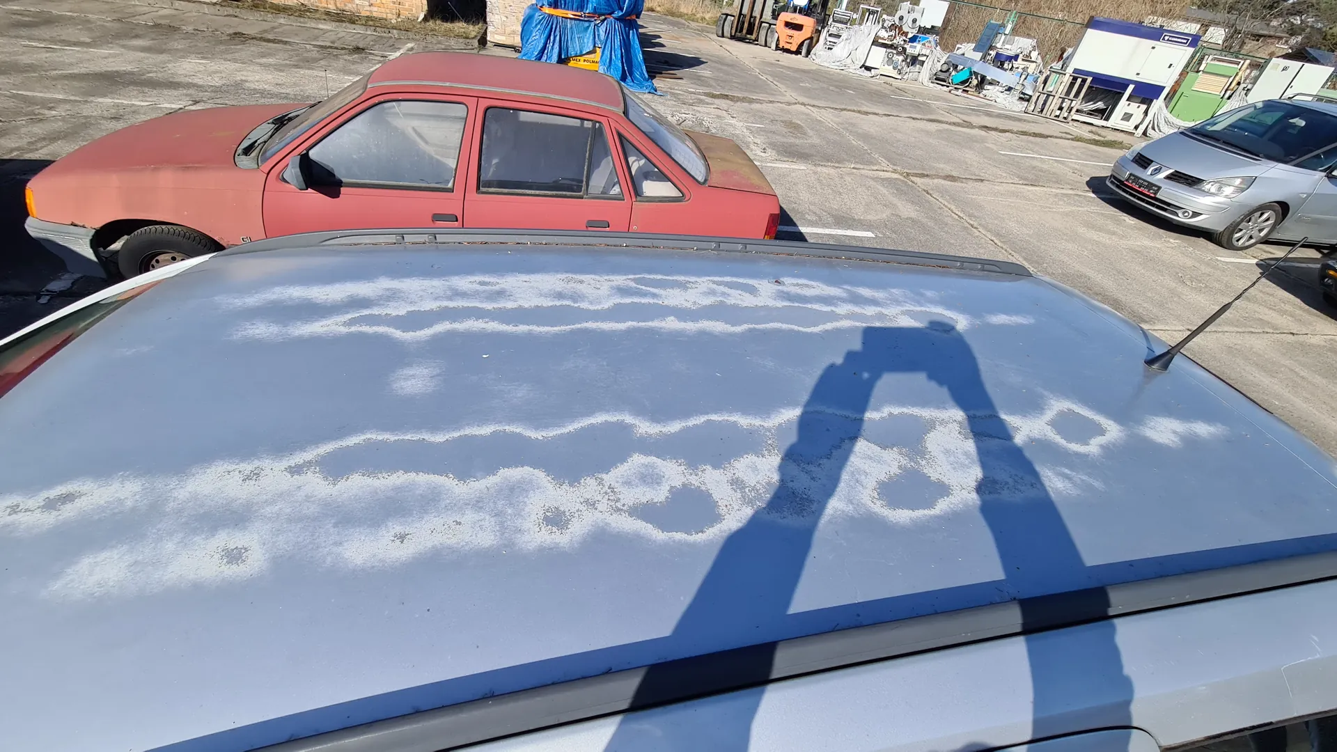 Dach jasnoszarego samochodu z białymi, rozmytymi śladami mrozu lub śniegu. Na dachu widoczny cień osoby trzymającej aparat lub telefon. W tle czerwony samochód, srebrny samochód, pomarańczowy wózek widłowy, niebieskie tkaniny oraz budynki przemysłowe na betonowym placu parkingowym.