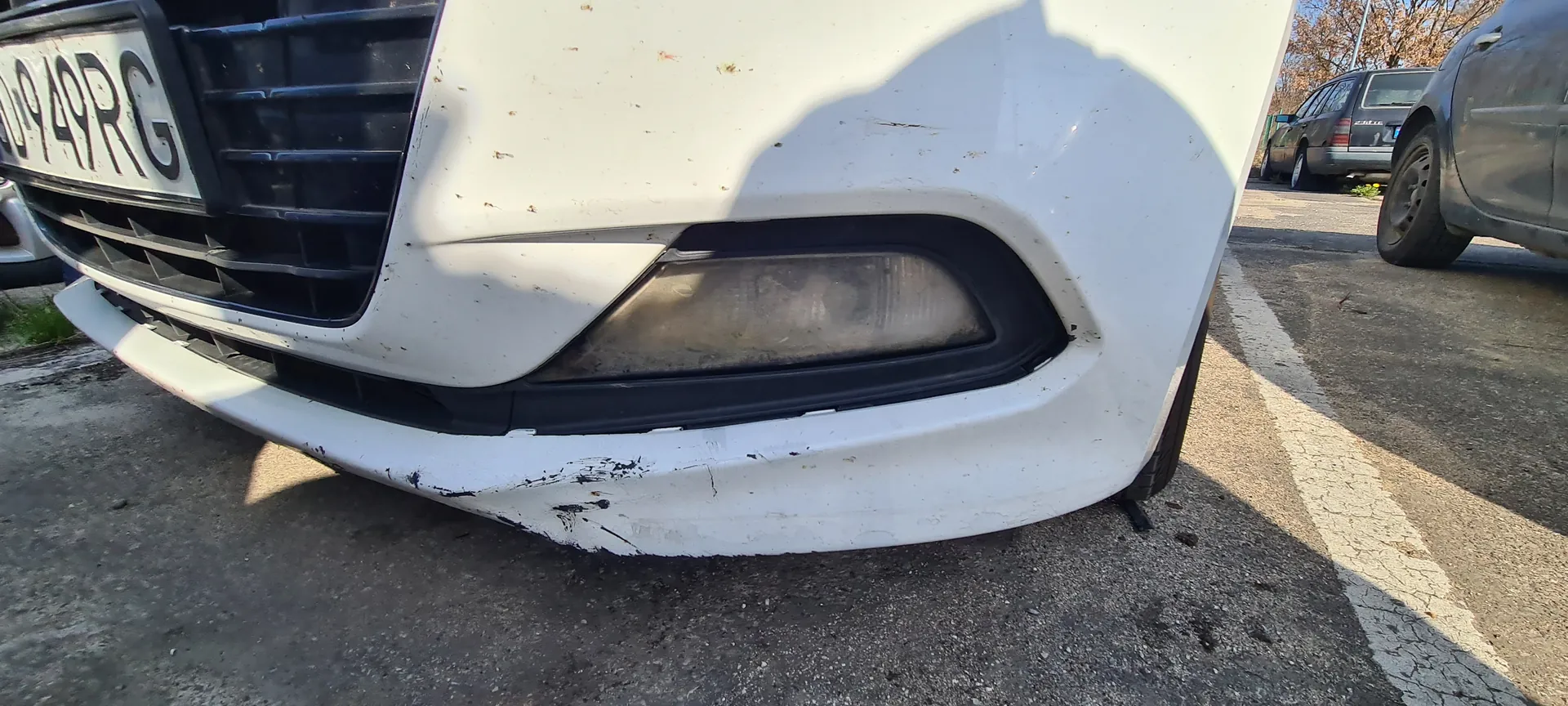Przód białego samochodu z widoczną tablicą rejestracyjną 0949RG. Na zderzaku widoczne są zadrapania i zabrudzenia. W tle inne zaparkowane samochody oraz asfaltowa powierzchnia parkingu.