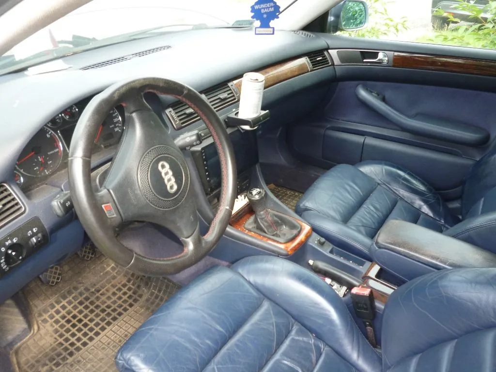 Wnętrze samochodu marki Audi z niebieską skórzaną tapicerką na fotelach. Widoczna jest czarna kierownica z logo Audi, deska rozdzielcza z drewnianymi elementami wykończenia, konsola środkowa z lewarkiem zmiany biegów oraz przedni pasażerski fotel. Na desce rozdzielczej, w uchwycie, znajduje się biały przedmiot w kształcie walca. W górnej części zdjęcia, przy lusterku wstecznym, widać niebieski odświeżacz powietrza z napisem 'WUNDER-BAUM'.