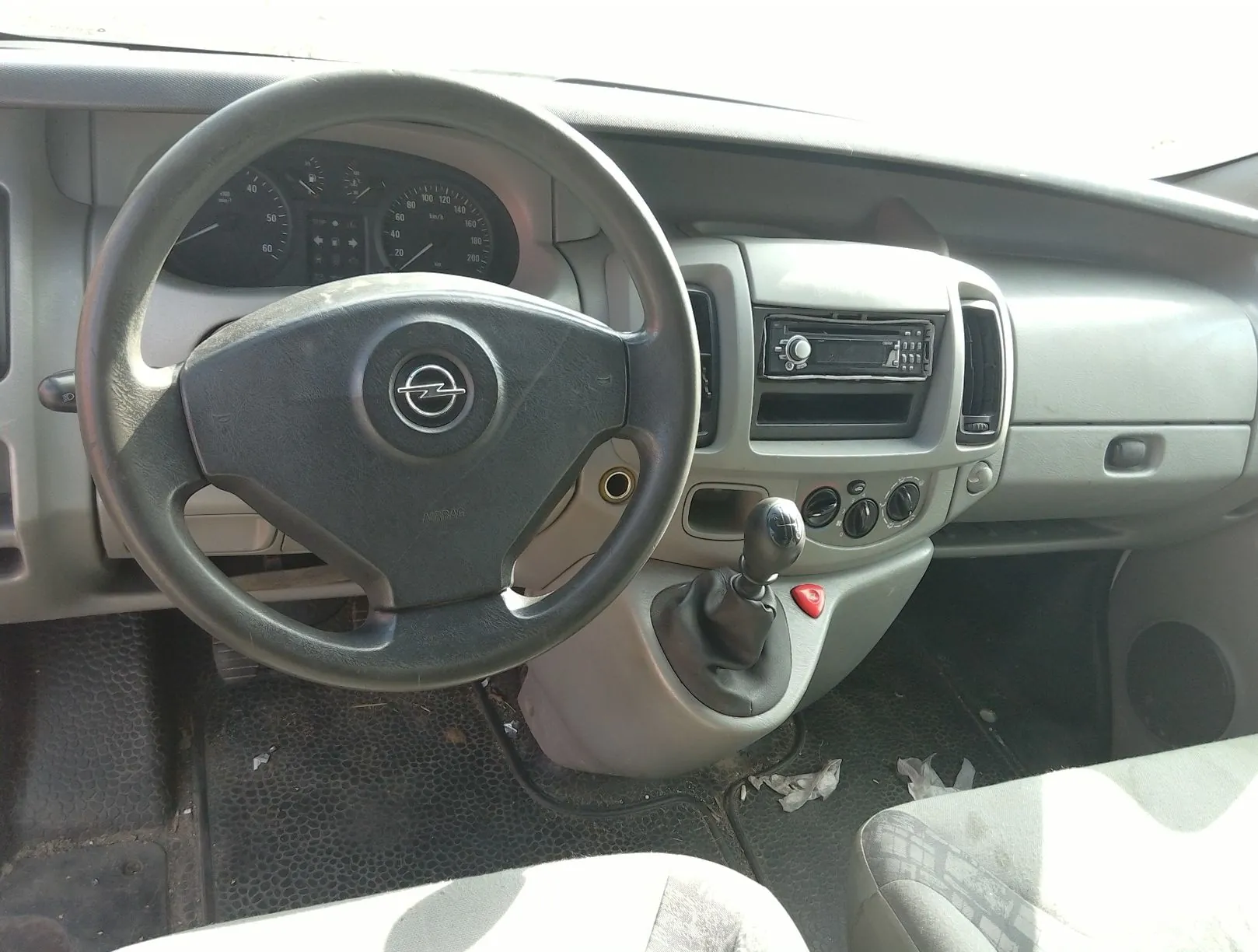 Wnętrze samochodu marki Opel, widok na deskę rozdzielczą i kierownicę. Widoczna jest czarna kierownica z logo Opela w centrum, zegary prędkościomierza i obrotomierza, radio w konsoli środkowej oraz drążek zmiany biegów. Tapicerka i elementy wnętrza są w kolorze szarym.