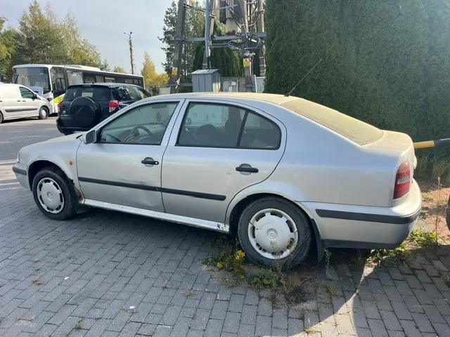 Srebrny samochód osobowy w nadwoziu sedan zaparkowany na kostce brukowej. Pojazd posiada widoczne uszkodzenia karoserii w postaci wgnieceń na drzwiach kierowcy oraz rdzy na dolnych krawędziach nadwozia. W tle widoczny jest czarny samochód, biały bus, biały autokar oraz zielone krzewy i drzewa.