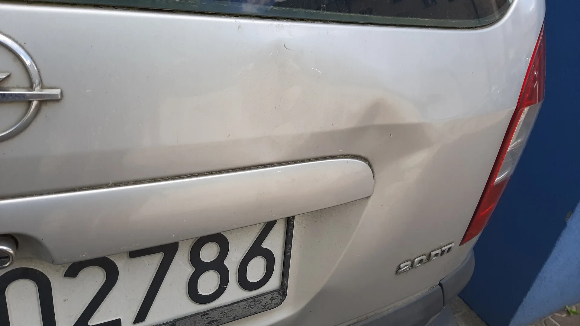 Zbliżenie na tylną część srebrnego samochodu. Widoczna biała tablica rejestracyjna z czarnymi cyframi 02786, logo marki po lewej stronie, napis 20DM po prawej stronie nad krawędzią bagażnika oraz czerwone światło tylnego reflektora. W tle fragment niebieskiej ściany i części ziemi. Samochód posiada drobne zabrudzenia i zadrapania na karoserii.