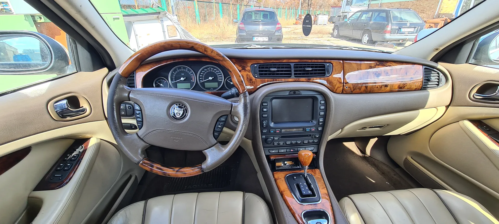 Wnętrze samochodu marki Jaguar z beżową skórzaną tapicerką. Widoczna kierownica z logo Jaguar i drewnianą obudową, deska rozdzielcza z analogowymi wskaźnikami prędkości i obrotomierza, centralna konsola z ekranem, przyciskami i dźwignią biegów z drewnianą rączką (oznaczenia 1-5, R). Na podłodze dywanik z napisem „JAGUAR”. Przez przednią szybę widoczne zaparkowane samochody, w tym ciemnoszary z tablicą rejestracyjną „GML 736M”, ogrodzenie i budynki. W lusterku bocznym odbicie zielonego pojazdu.