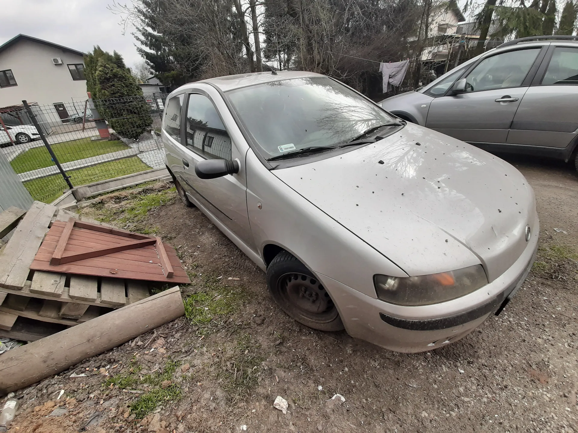 Srebrny samochód osobowy typu hatchback zaparkowany na podwórku z żwirem i trawą. Na masce widoczne są plamy i zabrudzenia. Obok znajduje się drugi srebrny samochód. W tle domy, ogrodzenie siatkowe, drzewa bez liści oraz pranie wiszące na sznurze. Przy lewym dolnym rogu obrazu leżą drewniane deski i belka. Na przedniej szybie pierwszego samochodu widoczny jest mały naklejony dokument. Niebo jest zachmurzone.