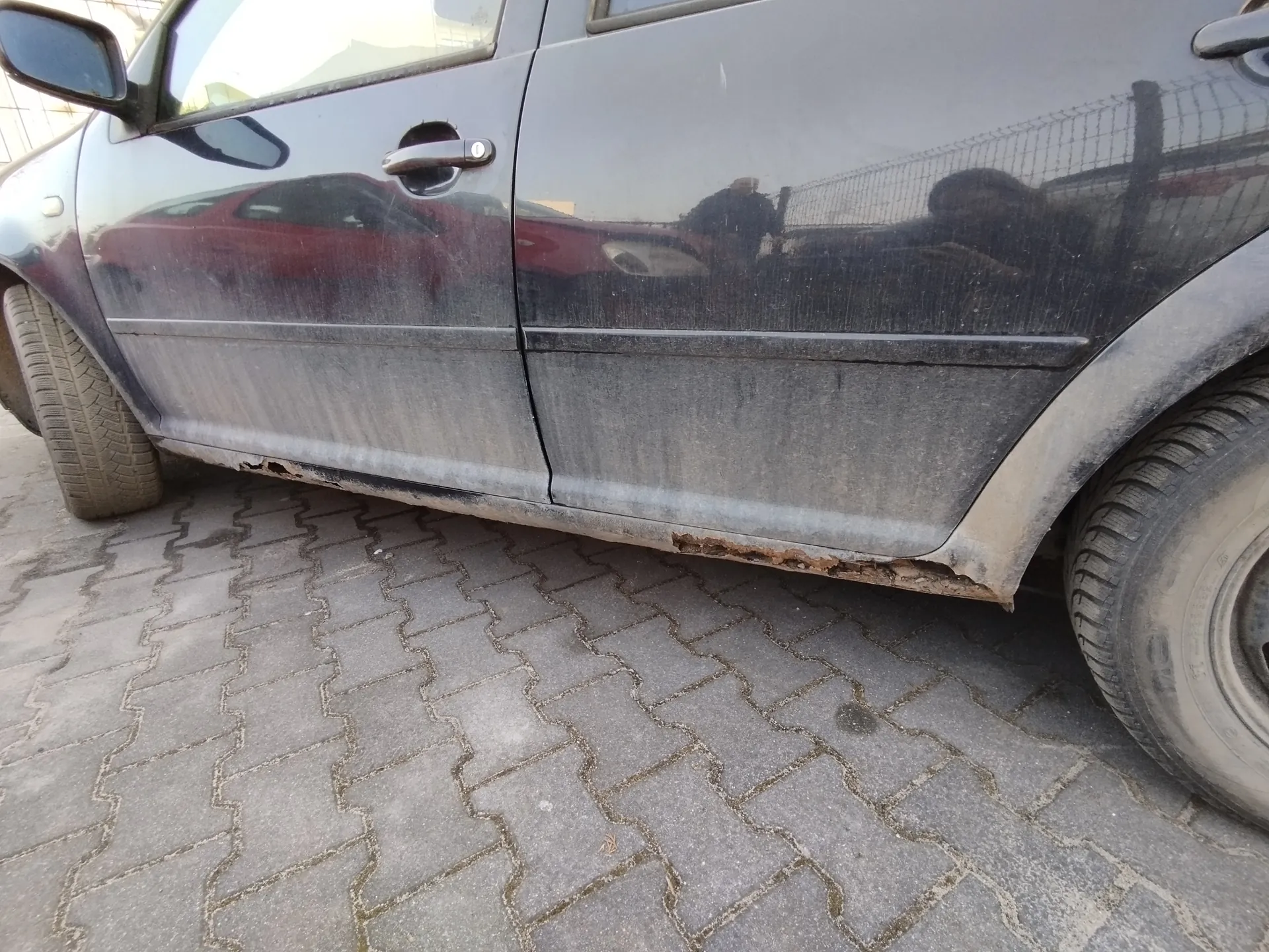 Boczny widok szarego samochodu z widocznymi śladami brudu i uszkodzeniami na dolnej krawędzi karoserii. Na tle chodnik z kostki brukowej oraz odbicie czerwonego samochodu i ogrodzenia w błyszczącej powierzchni drzwi.