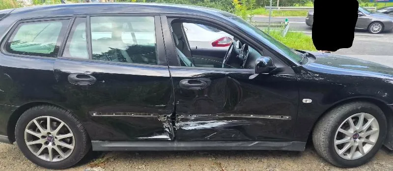 Czarny samochód typu kombi z widocznymi uszkodzeniami karoserii na prawym boku. Wgniecenia i zarysowania znajdują się na przednich i tylnych drzwiach pasażera, wzdłuż dolnej części nadwozia. Samochód stoi zaparkowany na poboczu obok trawy.
