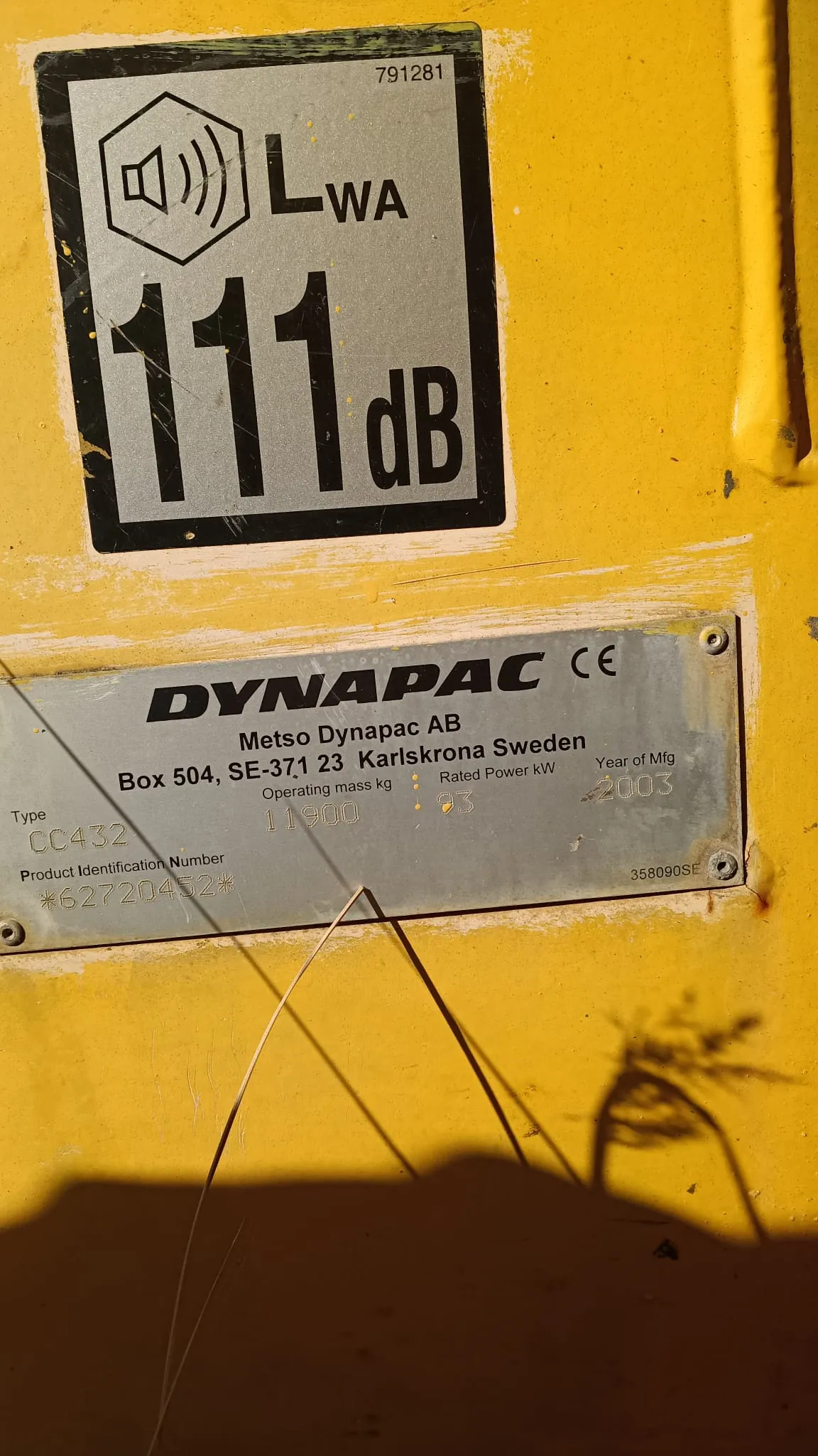 Zdjęcie dwóch tablic znamionowych na żółtej powierzchni maszyny. Górna tabliczka jest czarna z białym polem, zawiera ikonę głośnika, oznaczenie 'LWA' oraz napis '111 dB' i numer '791281'. Dolna tabliczka jest srebrna i zawiera tekst: 'DYNAPAC CE', 'Metso Dynapac AB', 'Box 504, SE-371 23 Karlskrona Sweden'. Poniżej znajdują się dane techniczne: 'Operating mass kg 11900', 'Rated Power kW 93', 'Year of Mfg 2003', 'Type CC432', 'Product Identification Number *62720432*' oraz numer '358090SE'.