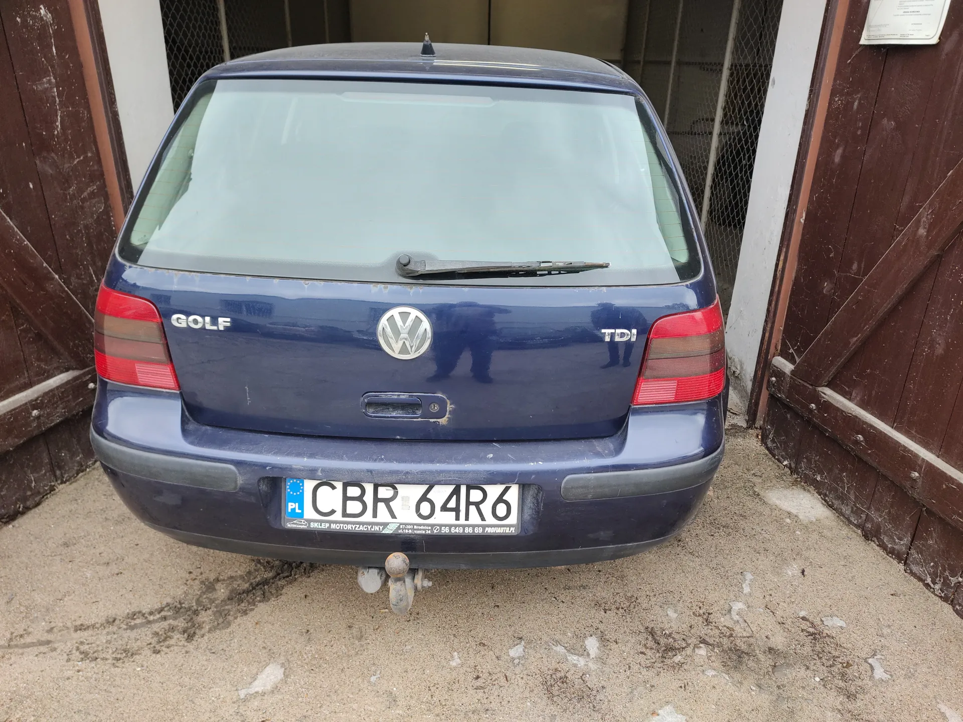 Tył niebieskiego samochodu Volkswagen Golf TDI zaparkowanego między dwiema brązowymi drewnianymi bramami. Na bagażniku widoczne napisy "GOLF" po lewej stronie i "TDI" po prawej stronie, a w centrum logo Volkswagena. Tablica rejestracyjna z białym tłem i czarnymi literami "CBR 64R6", z niebieskim pasem po lewej stronie z napisem "PL" oraz dodatkowym tekstem poniżej tablicy. Samochód stoi na betonowej powierzchni z widocznymi plamami brudu. W tle siatka metalowa i fragmenty budynku.