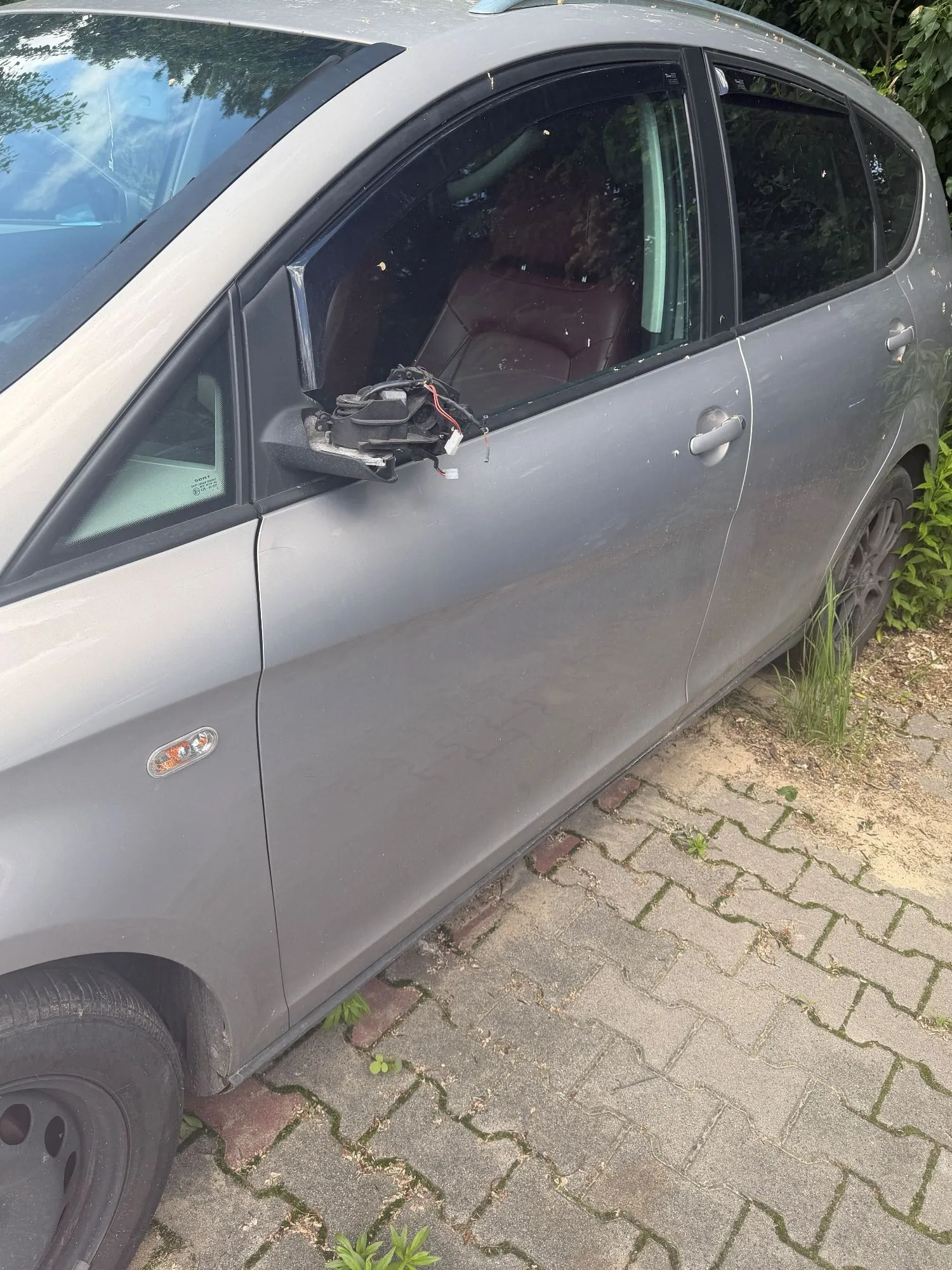 Zbliżenie na prawy bok srebrnego samochodu. Przednia szyba boczna jest rozbita, a w miejscu lusterka zewnętrznego widoczne są wyrwane elementy plastikowe oraz zwisające przewody elektryczne.
