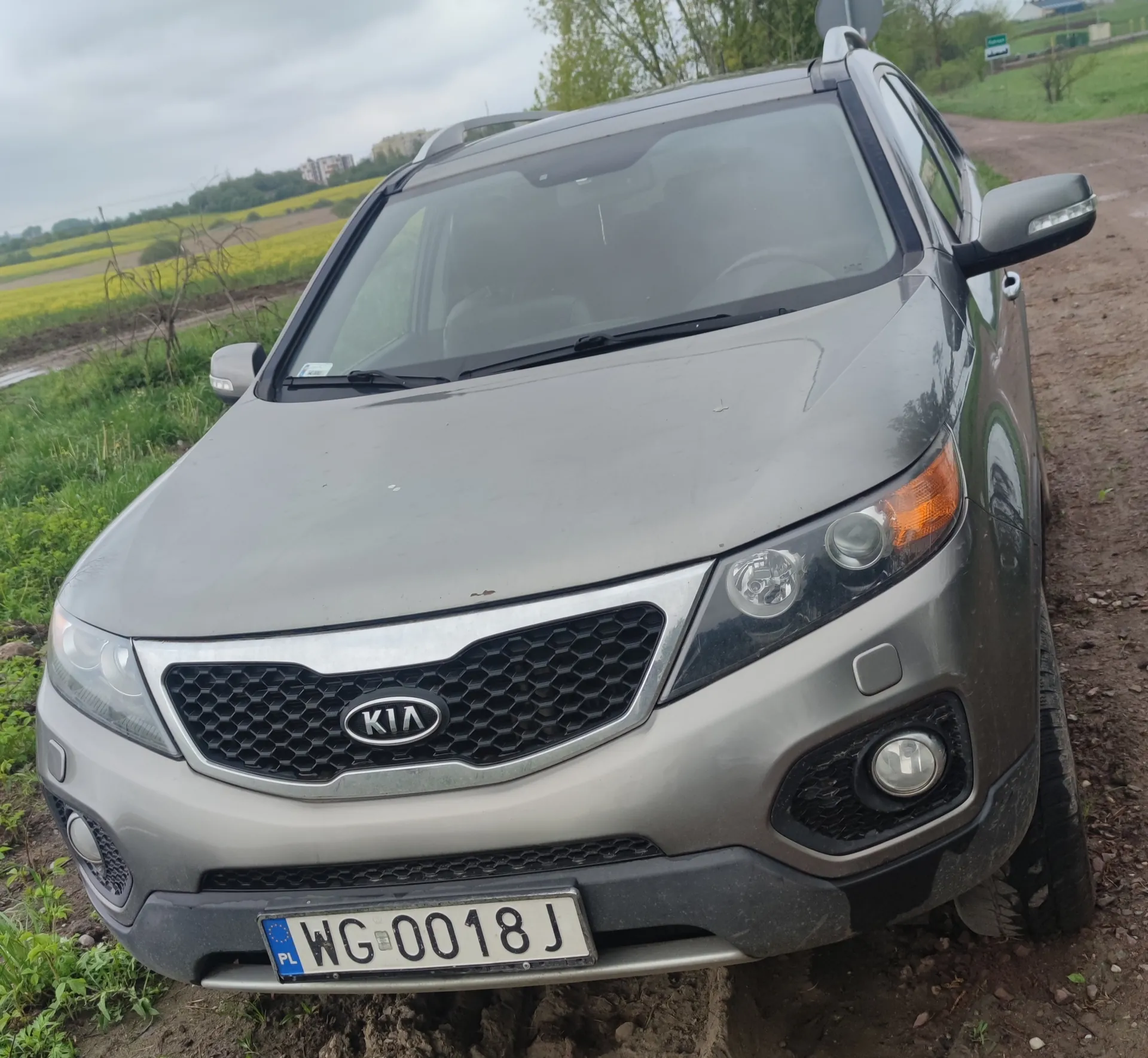 Przedni widok szarego SUV-a marki Kia z tablicą rejestracyjną WG-0018 J (PL) na błotnistej drodze. W tle pola z żółtymi uprawami, zielona trawa, drzewa i chmurne niebo. Widoczne są również budynki w oddali oraz znak drogowy.