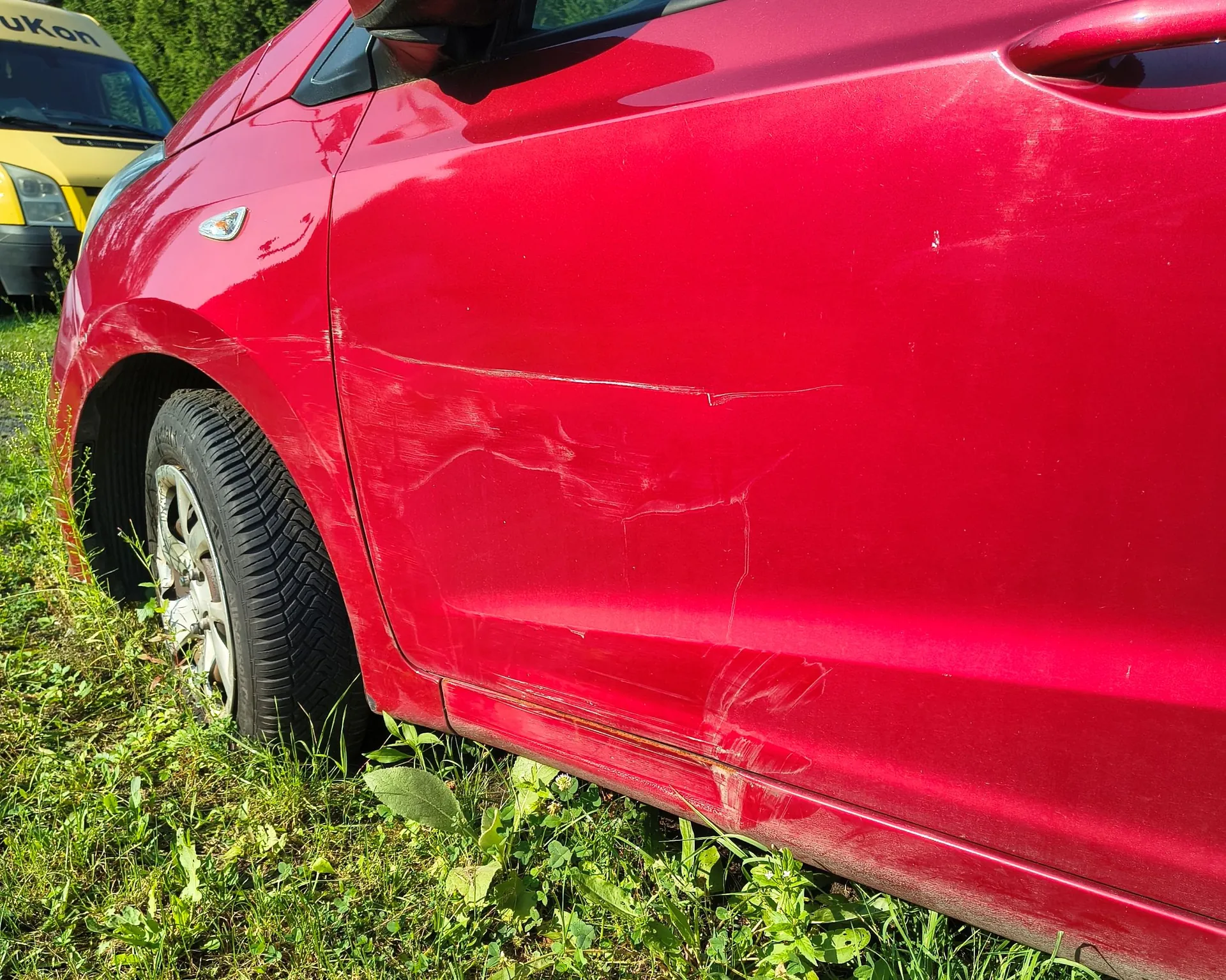 Zbliżenie na prawy bok czerwonego samochodu, pokazujące liczne zarysowania i wgniecenia na drzwiach pasażera oraz tylnym błotniku. Auto stoi na trawie, w tle widoczny jest fragment żółtego pojazdu z napisem 'ukOn'.