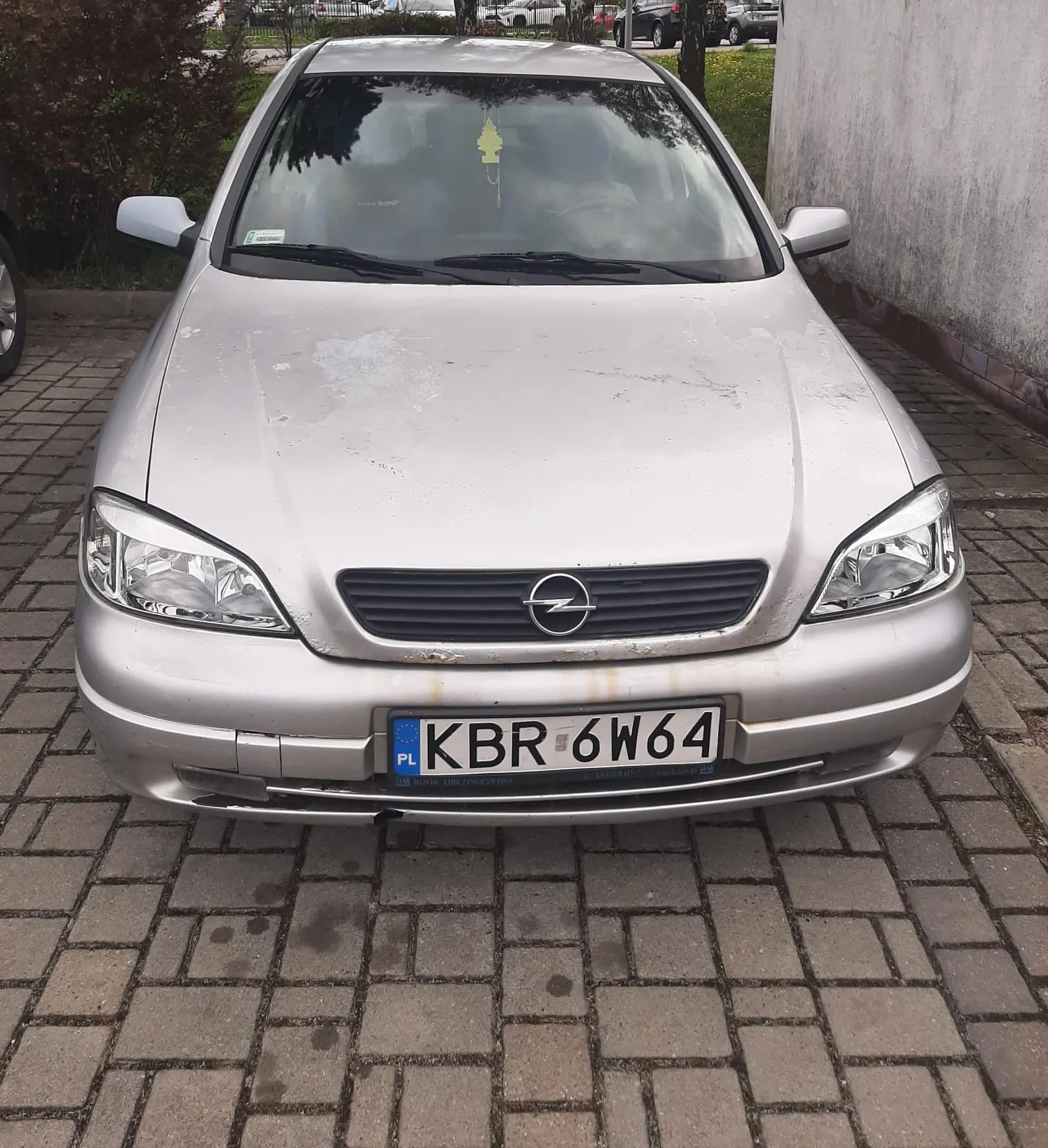 Srebrny samochód marki Opel zaparkowany na brukowanej powierzchni. Widoczna przód pojazdu z logo Opel na atrapie chłodnicy. Tablica rejestracyjna z polskim kodem PL i numerem KBR 6W64. Na przedniej szybie wiszący żółty odświeżacz powietrza. Tło: ściana po prawej stronie, krzewy po lewej oraz inne zaparkowane samochody w oddali. Samochód pokryty jest plamami brudu lub wody na masce.
