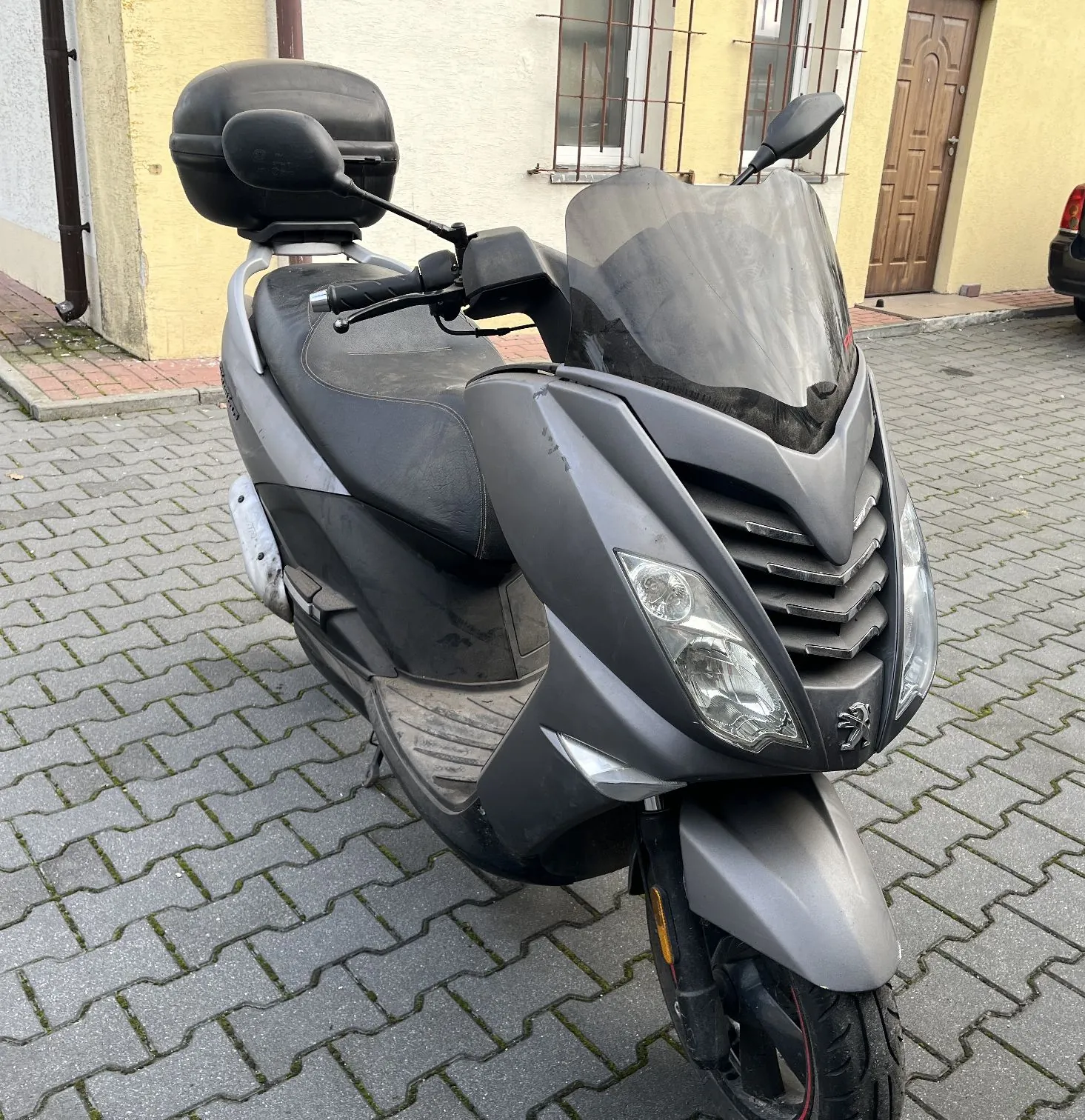 Srebrny skuter marki Peugeot zaparkowany na kostce brukowej przed jasnym budynkiem. Pojazd posiada czarną kanapę, przednią szybę ochronną oraz czarny kufer zamontowany z tyłu.