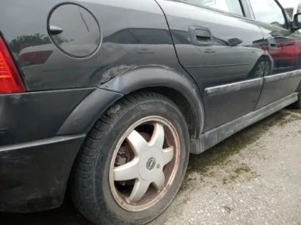 Czarny samochód widziany od tyłu z widocznym uszkodzeniem na tylnym błotniku. Srebrna felga z logo w centrum, czerwone światło tylnego reflektora. Samochód zaparkowany na nawierzchni z drobnych kamieni i plam mchu. W tle widoczne inne pojazdy, w tym czerwony samochód.