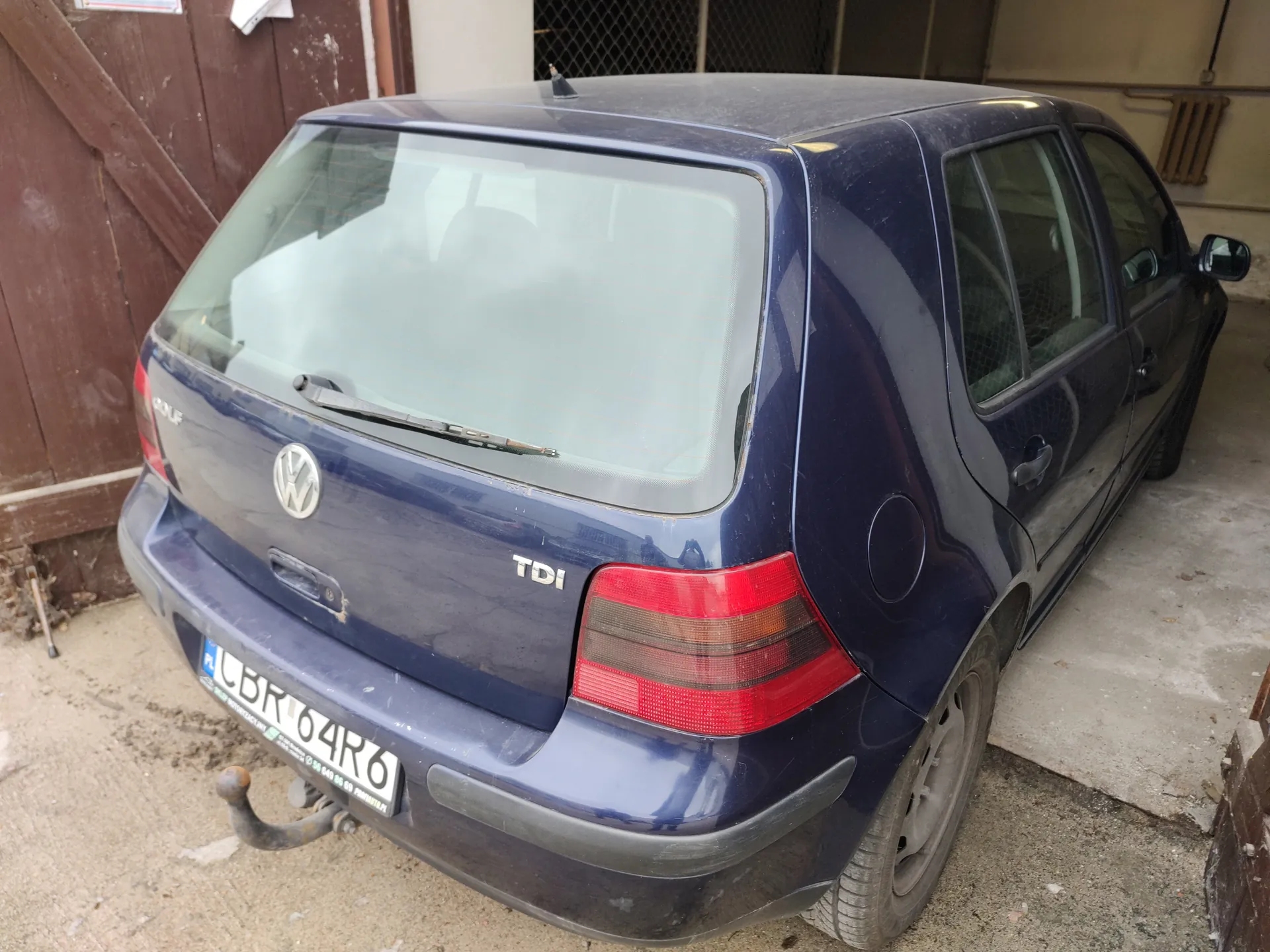 Ciemnoniebieski samochód osobowy marki Volkswagen Golf TDI, widok od tyłu. Na bagażniku widoczny emblemat VW oraz napis TDI. Tablica rejestracyjna z polskim kodem PL i numerem CBR 64R6. Samochód stoi na betonowej powierzchni przed brązowymi drzwiami garażowymi. W tle widoczne siatki i fragment budynku. Na tylnej szybie umieszczony wycieraczka. Przy zderzaku tylnym zamontowany hak holowniczy.