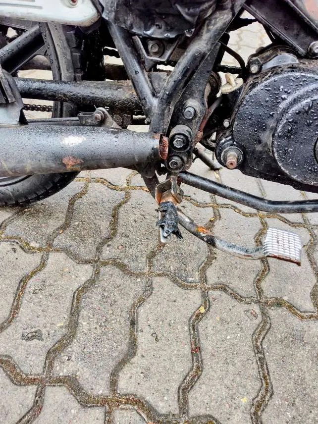 Zbliżenie na dolną część motocykla, w tym rurę wydechową z widocznymi plamami rdzy, podnóżek z teksturą, elementy ramy i silnika pokryte kroplami wody. Tło stanowi brukowana powierzchnia z kamiennymi płytami o nieregularnym kształcie, z zielonymi zabrudzeniami w szwach.