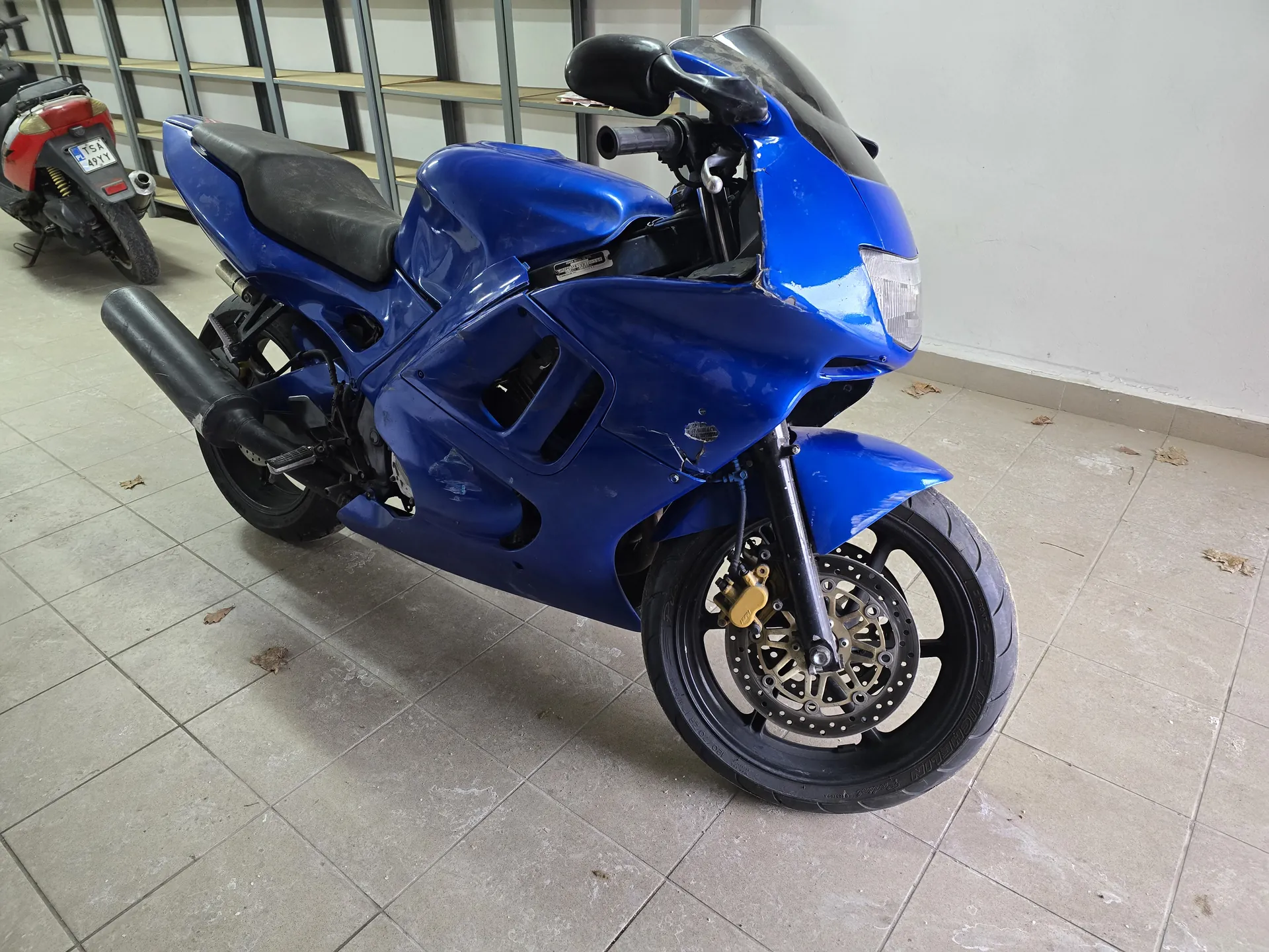 Niebieski motocykl sportowy z czarnym siedzeniem i widocznymi uszkodzeniami na karoserii, stojący na podłodze z szarymi płytkami. W tle czerwony skuter z tablicą rejestracyjną T54 491Y oraz metalowe półki. Na podłodze rozrzucone są suche liście.