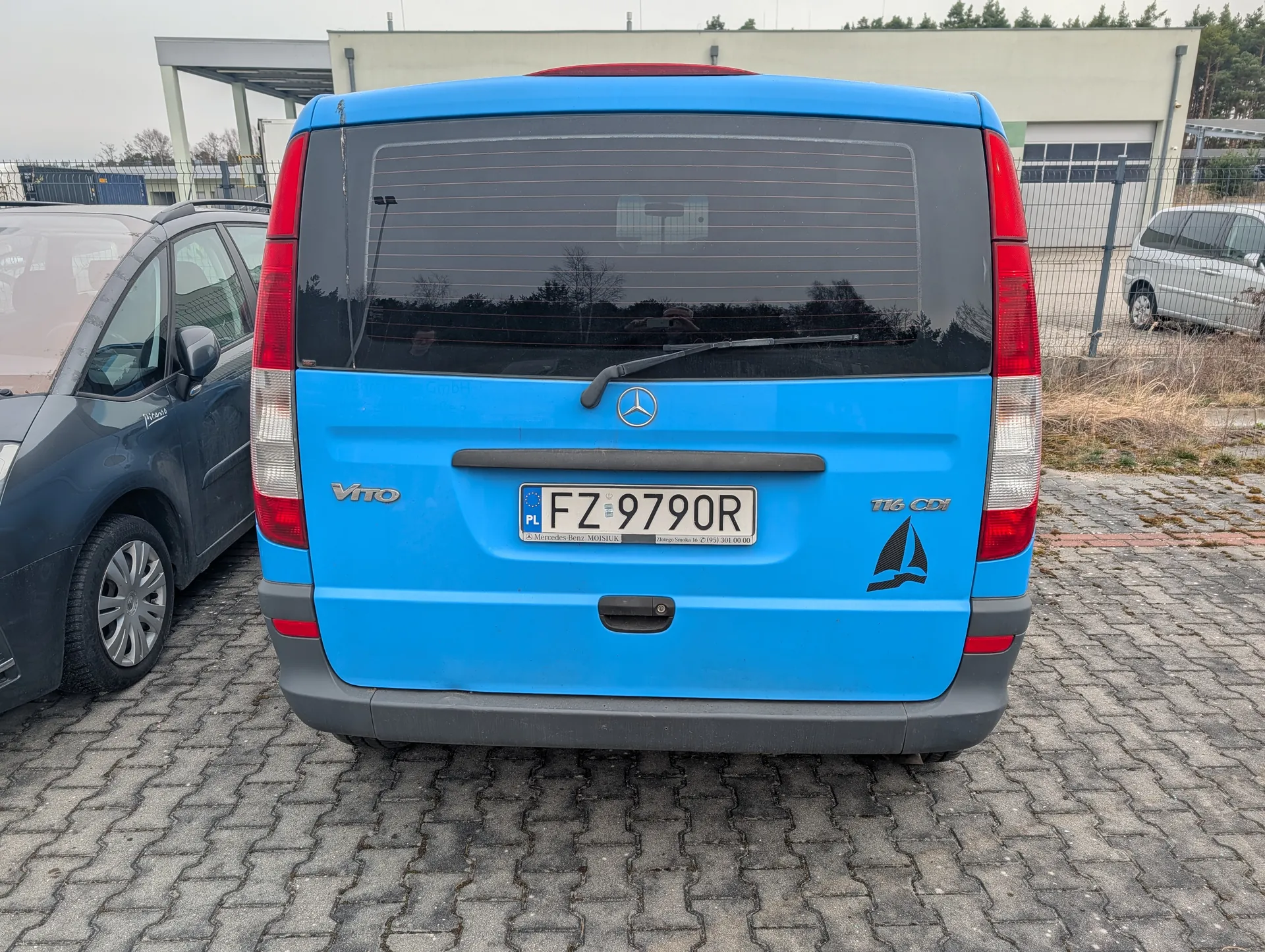 Z tyłu niebieski samochód dostawczy Mercedes-Benz Vito z tablicą rejestracyjną PL FZ 9790R. Na tylnej szybie widoczny wycieraczka, a na boku napisy 'Vito' i '116 CDI' oraz naklejka z czarną żaglówką. Po lewej stronie ciemnoszary samochód osobowy, po prawej srebrny samochód. Tło: budynek przemysłowy z bramą garażową, ogrodzenie i suche trawy na brukowanym placu parkingowym. Niebo jest pochmurne.