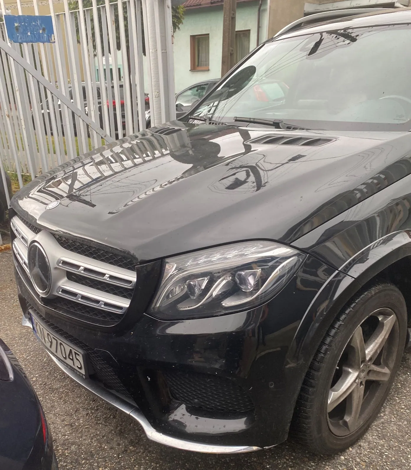 Zbliżenie na przód czarnego samochodu marki Mercedes-Benz. Widoczna jest maska, reflektor prawy, grill z dużym logo marki oraz fragment tablicy rejestracyjnej z numerem 97045. Samochód zaparkowany jest obok metalowego ogrodzenia z pionowych prętów.