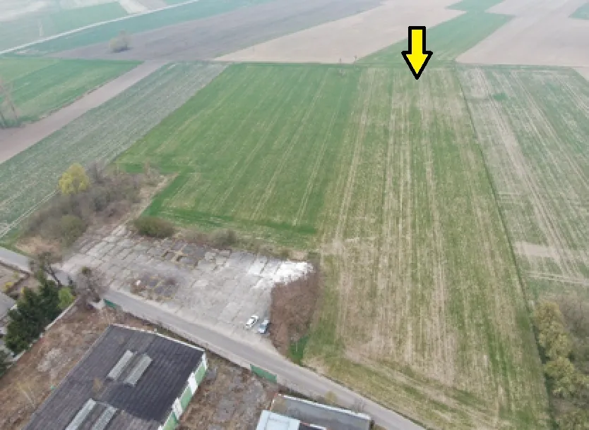 Zdjęcie lotnicze przedstawiające obszar rolniczy z polami w różnych odcieniach zieleni i brązu. W lewym dolnym rogu widoczny jest betonowy plac oraz fragmenty dachów budynków przemysłowych. W górnej części obrazu znajduje się żółta strzałka skierowana w dół, wskazująca na konkretny punkt na zielonym polu.