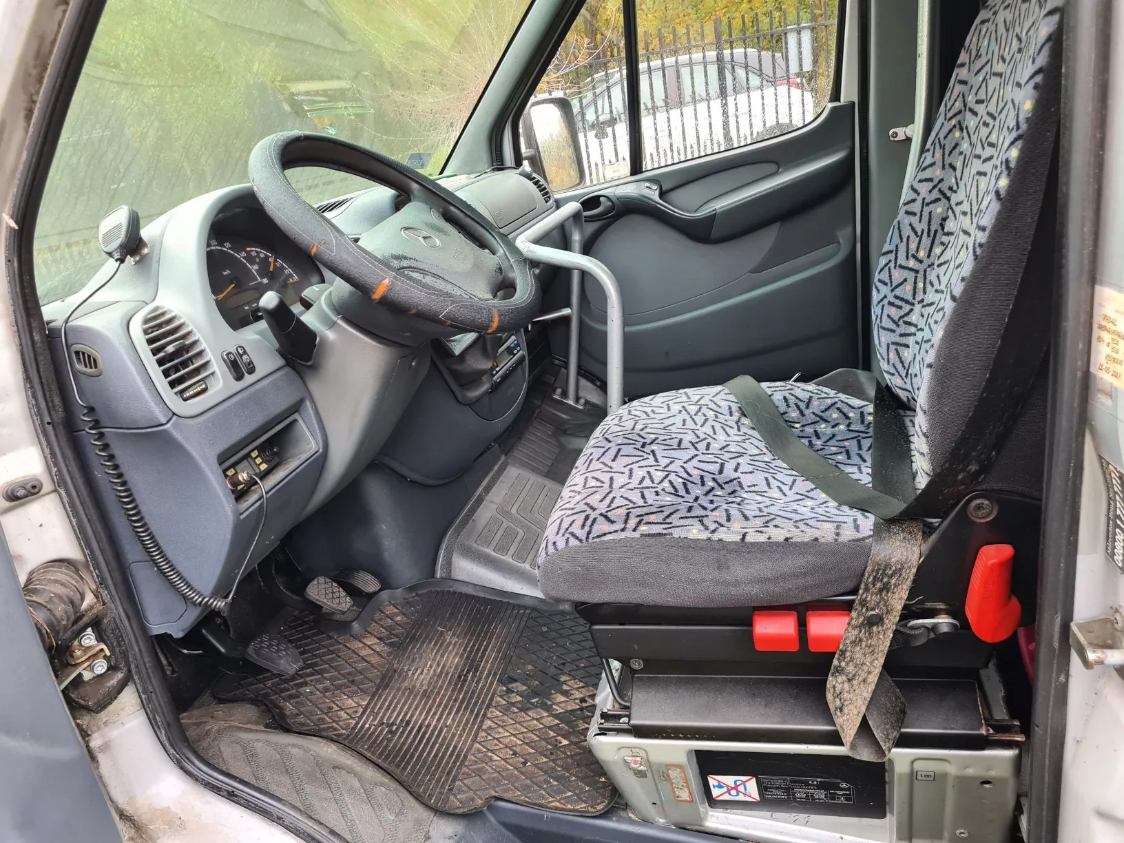 Wnętrze pojazdu widziane z otwartego drzwi kierowcy. Kierownica z czarnym obiciem i pomarańczowymi akcentami, deska rozdzielcza z licznikami, przyciskami i wentylacją. Siedzenie kierowcy z wzorzystym obiciem w szaro-czarne kształty, z zapiętym pasem bezpieczeństwa. Czerwone przyciski na podstawie siedzenia. Brudne dywaniki na podłodze. Poza pojazdem widoczny płot, drzewa i fragment innego samochodu. Na panelu pod siedzeniem widoczne etykiety z symbolami i niewyraźnym tekstem.