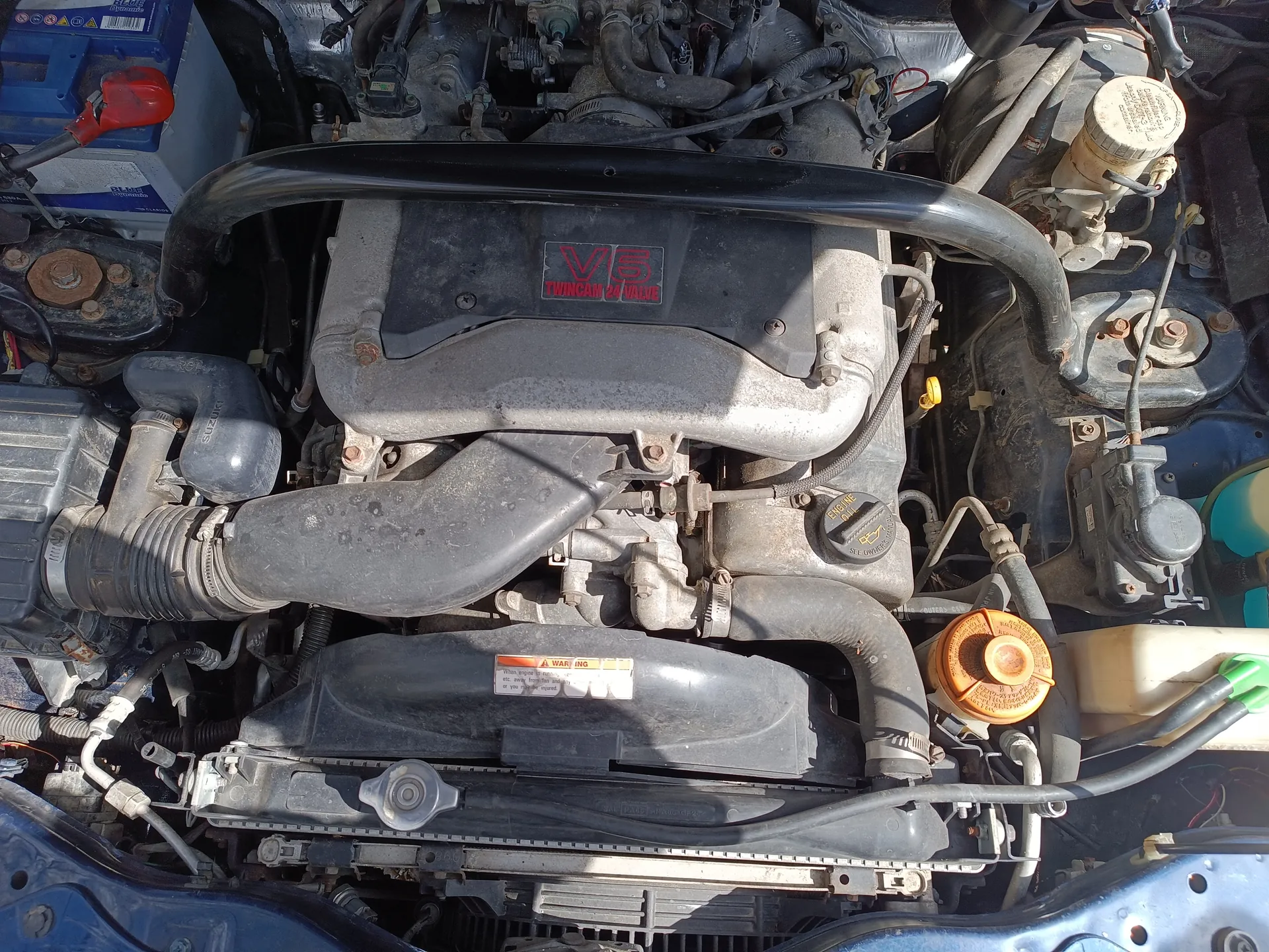 Zbliżenie na silnik samochodowy z widoczną pokrywą z napisem "V6 TWINCAM 24 VALVE" w czerwonych literach. Po lewej stronie niebieski akumulator z czerwoną nakrętką, różne przewody, węże i elementy mechaniczne. W dolnej części czarna osłona z białą etykietą ostrzegawczą. Obecny pomarańczowy korek zbiornika chłodniczego oraz zbiornik z zielonym nakrętką po prawej stronie. Silnik pokryty jest kurzem, widoczne ślady eksploatacji.