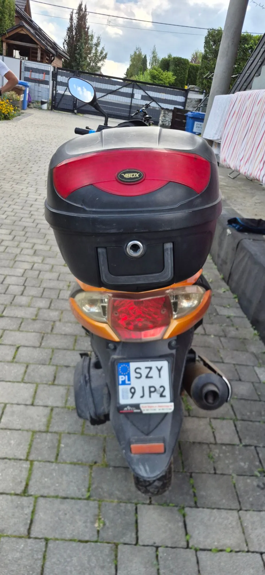 Widok z tyłu na pomarańczowy skuter zaparkowany na kostce brukowej. Na tylnym bagażniku zamontowany jest czarny kufer z czerwonym elementem dekoracyjnym i napisem 'VBOX'. Skuter posiada polską tablicę rejestracyjną o numerze SZY 9JP2.