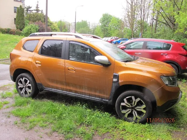 Pomarańczowy SUV zaparkowany na trawie, obok czerwonego samochodu. W tle widoczne są inne pojazdy, budynki, drzewa i krzewy. Na dole obrazu znajduje się data 11/05/2026. Samochody stoją na nierównym terenie z zieloną trawą i błotem.