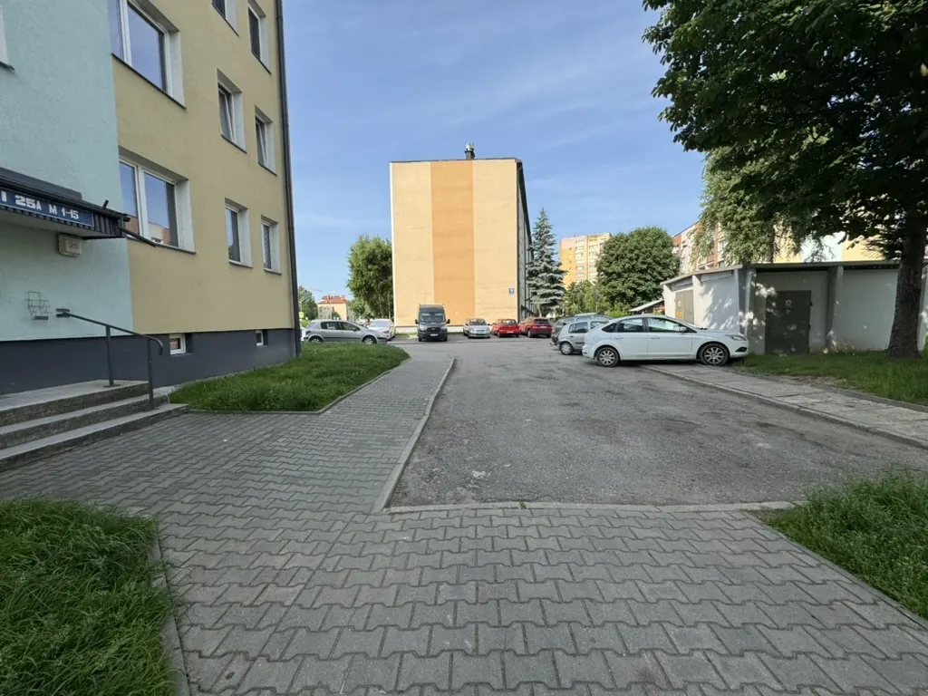 Ścieżka z bruku z rampą dla niepełnosprawnych biegnie między budynkami. Po lewej budynek z tablicą „25A M 1-15”, po prawej zaparkowane samochody. W tle brązowo-żółty blok mieszkalny, drzewa i jasne niebo. Trawniki po obu stronach.