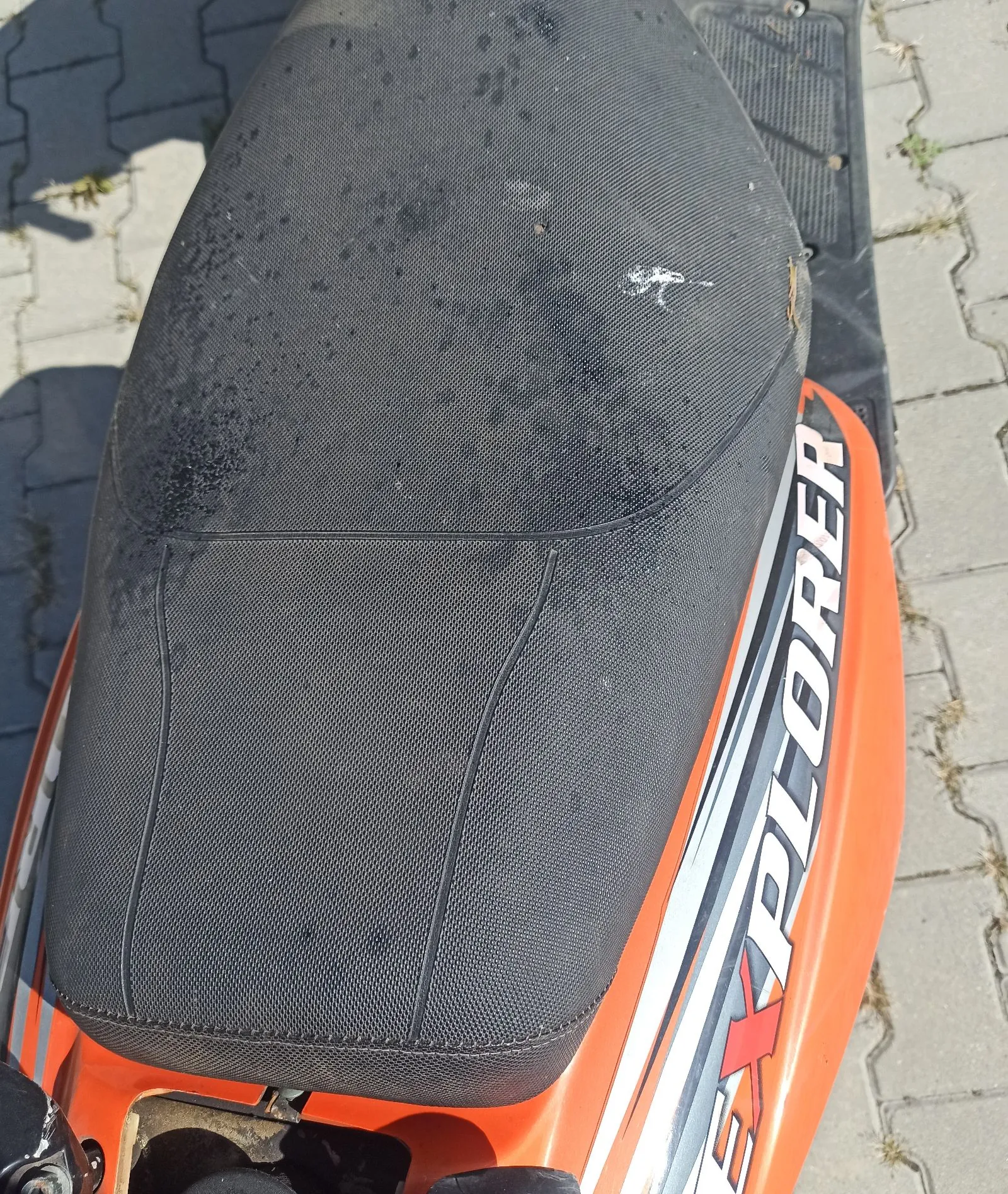 Widok z góry na czarne, teksturowane siedzenie pomarańczowego skutera. Na boku skutera widnieje biały napis z czarnym obrysem: 'EXPLORER'. Pojazd stoi na kostce brukowej.