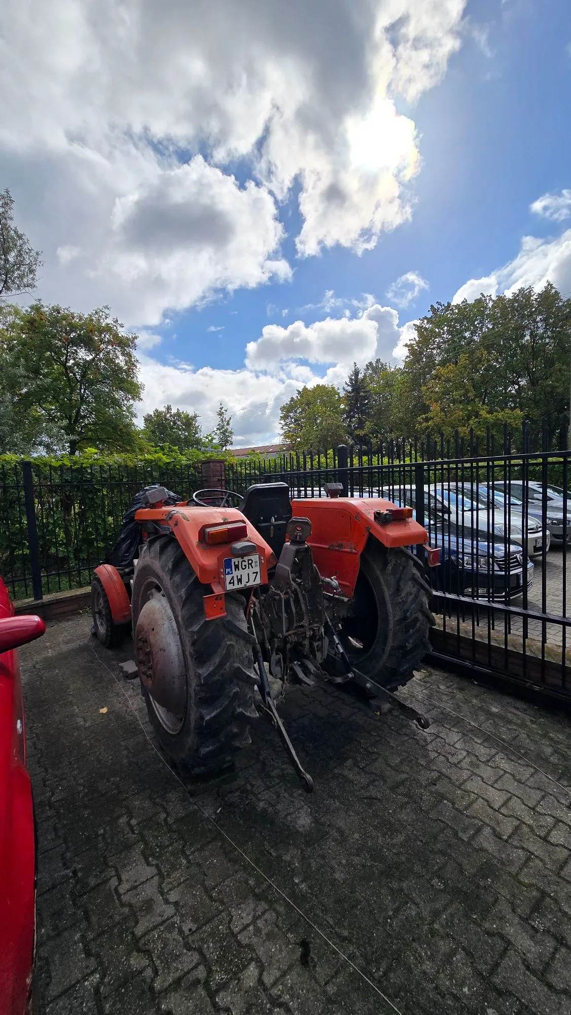 Pomarańczowy traktor zaparkowany na kostce brukowej przed czarnym metalowym ogrodzeniem. Traktor ma duże tylne koła z wyraźnym bieżnikiem i mniejsze przednie koła. Na tylnej części pojazdu widoczna jest tablica rejestracyjna o treści 'WGR 4WJ7'. W tle widać zielone drzewa oraz zaparkowane samochody za ogrodzeniem, a nad nimi niebo z białymi chmurami i słońcem.
