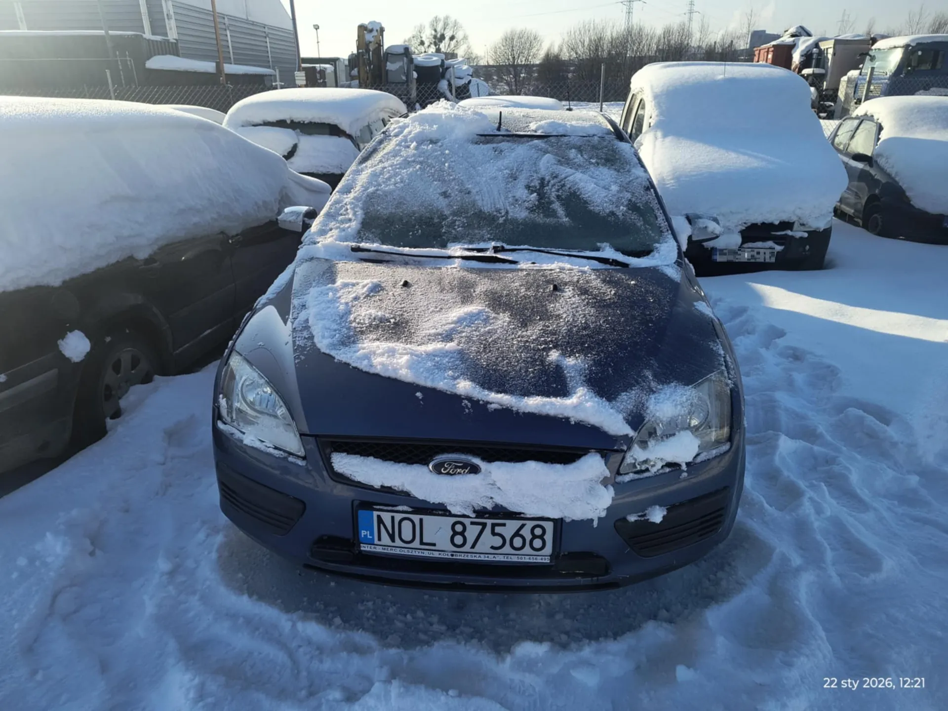 Zdjęcie przedstawia parking pokryty śniegiem z kilkoma samochodami. W centrum kadru znajduje się ciemnoniebieski samochód marki Ford z widocznym logo na masce. Na masce i przedniej szybie leży śnieg. Tablica rejestracyjna z białym tłem i czarnym napisem: PL NOL 87568. W tle widoczne są inne samochody zakryte śniegiem, budynki przemysłowe, ogrodzenie i drzewa bez liści. W prawym dolnym rogu zdjęcia znajduje się znacznik czasu: 22 sty 2026, 12:21. Słońce świeci, rzucając cienie na śnieg.