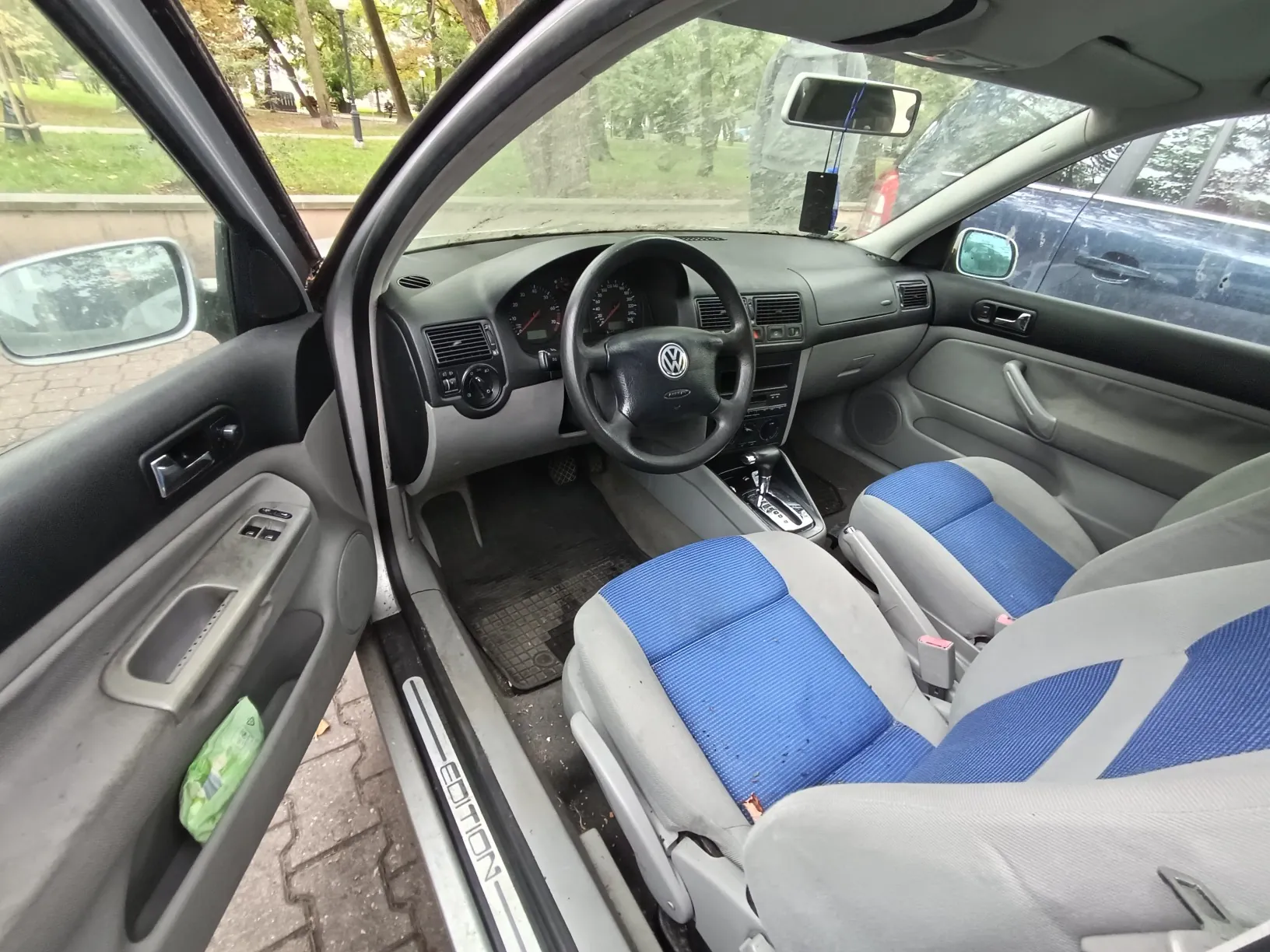 Wnętrze samochodu marki Volkswagen widziane z perspektywy otwartych przednich drzwi kierowcy. Widoczna jest szara deska rozdzielcza, czarna kierownica z logo VW, wskaźniki prędkości i obrotów oraz dwa przednie fotele z szaro-niebieską tapicerką. Na progu samochodu widnieje napis 'EDITION'. Przez przednią szybę i okna widać zieleń drzew w tle.