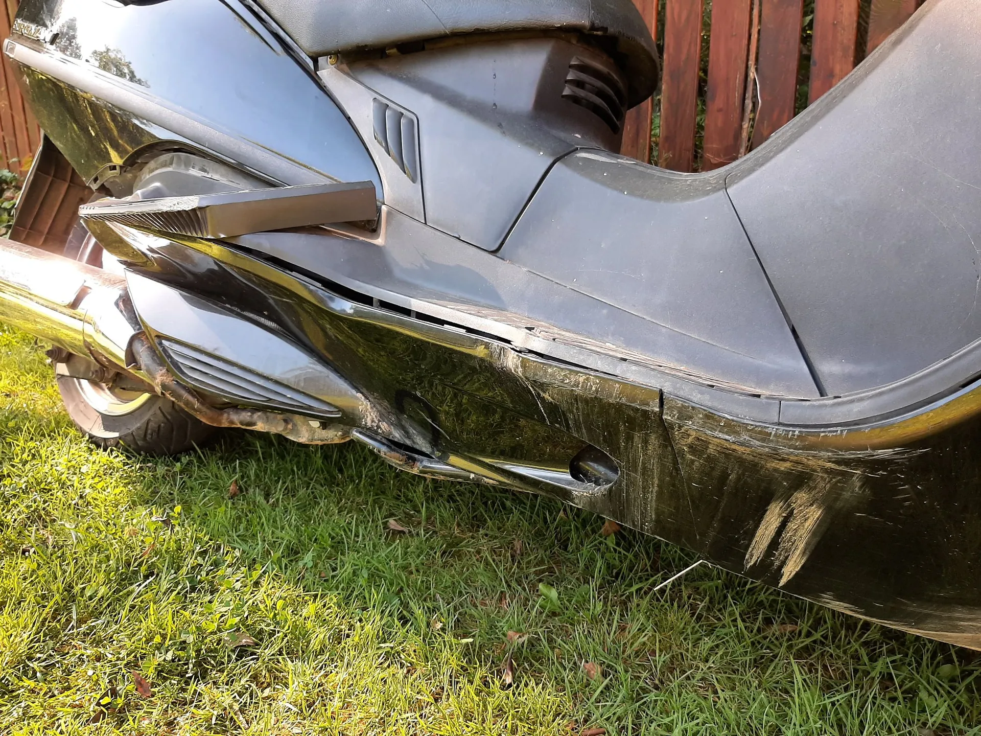 Zbliżenie na uszkodzoną, czarną obudowę plastikową skutera. Widoczne są głębokie zarysowania, przetarcia oraz pęknięcie i odkształcenie panelu bocznego w dolnej części pojazdu. W tle widać fragment koła, rurę wydechową oraz zieloną trawę i drewniany płot.