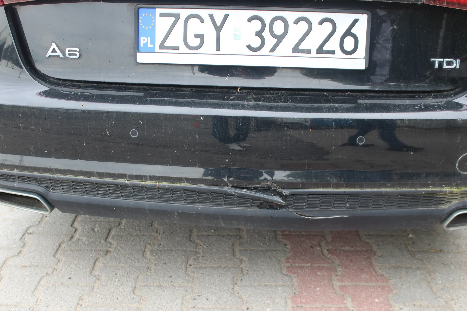 Zbliżenie na tylny zderzak czarnego samochodu marki Audi A6 TDI. Widoczna jest polska tablica rejestracyjna o numerze ZGY 39226. Dolna część zderzaka, w obszarze czarnej kratki, jest wgnieciona i uszkodzona. Samochód stoi na kostce brukowej.