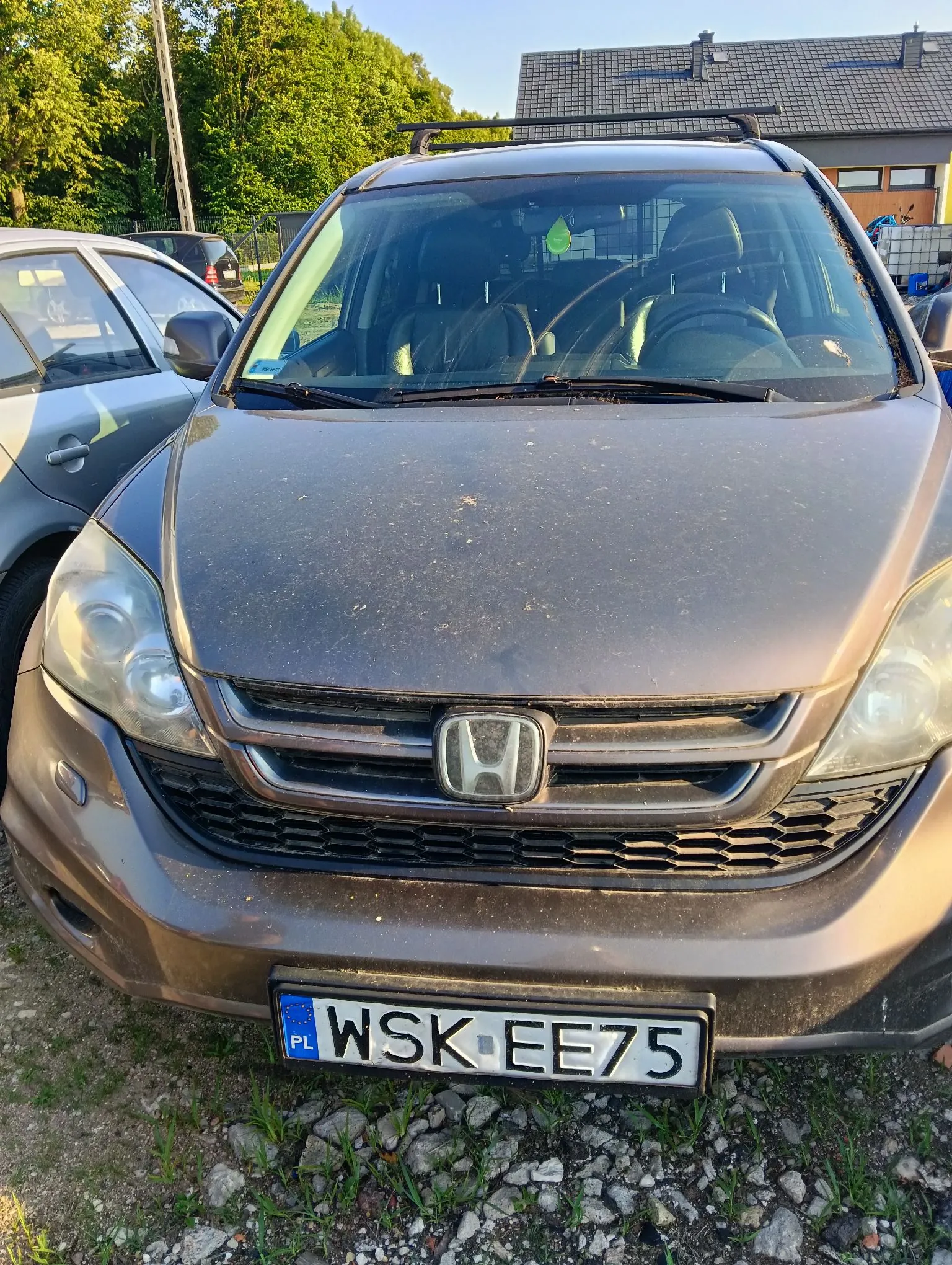 Zdjęcie przodu brązowego samochodu marki Honda z zamontowanym bagażnikiem dachowym. Pojazd jest zakurzony i zaparkowany na żwirowym podłożu obok innego, srebrnego samochodu. Widoczna jest tablica rejestracyjna o treści WSK EE75 z oznaczeniem PL. W tle widać zielone drzewa oraz fragment budynku z ciemnym dachem.