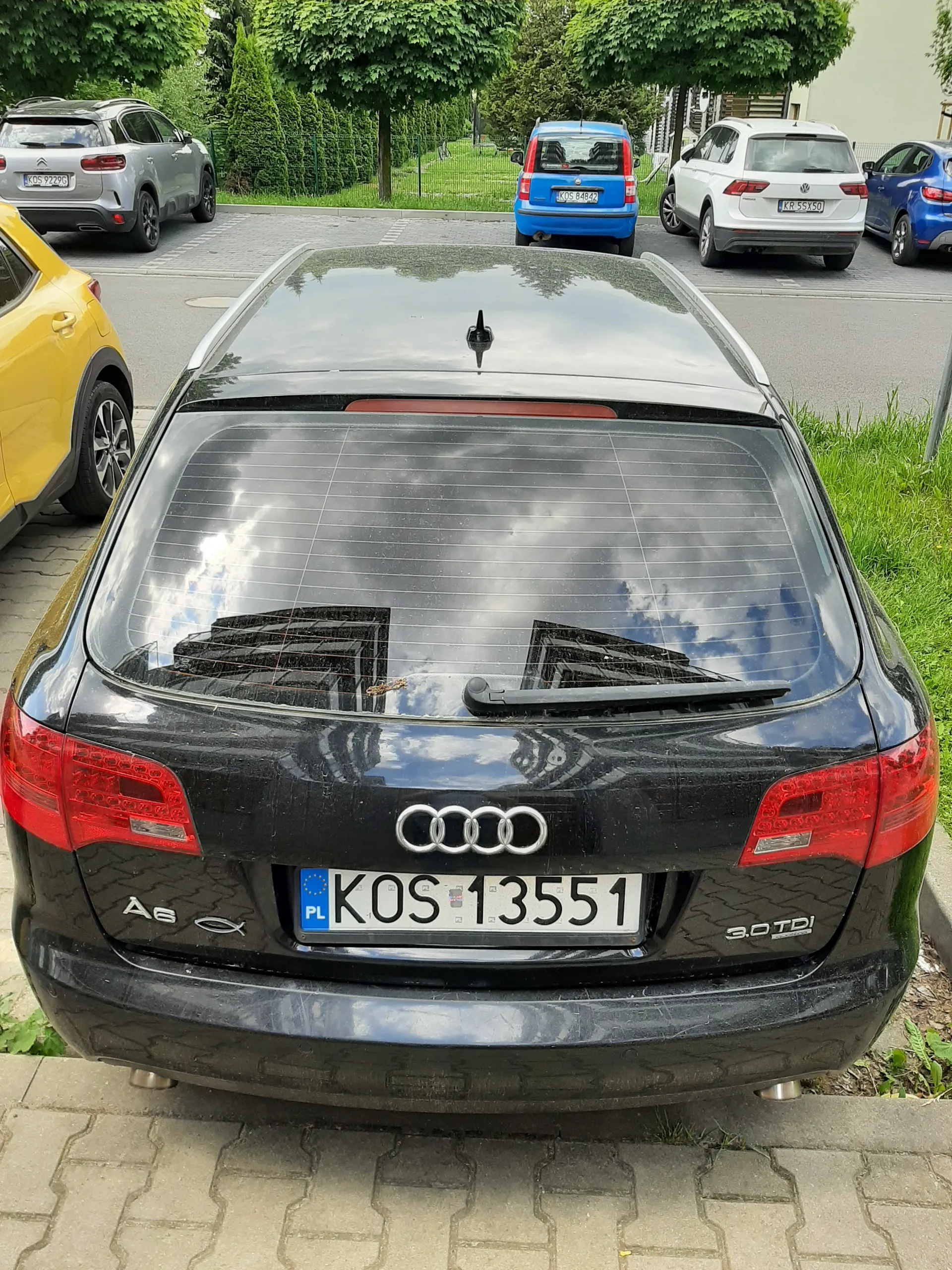 Z tyłu czarnego samochodu marki Audi A6 z widocznym logo, napisem '3.0 TDI' oraz 'A6'. Tablica rejestracyjna KOS 13551 z oznaczeniem 'PL' i symbolem Unii Europejskiej. W tle inne zaparkowane samochody: biały, niebieski, żółty i srebrny. Po lewej stronie zielone krzewy i trawa, po prawej chodnik z kostki. Na szybie tylnej odbicie budynków i nieba z chmurami.