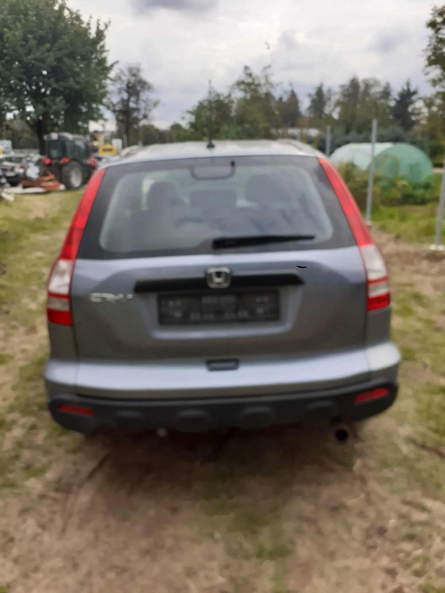 Tylny widok srebrnego samochodu marki Honda w nadwoziu kombi, zaparkowanego na trawiastym terenie. Samochód ma zamaskowaną tablicę rejestracyjną. W tle widoczne są drzewa, zielone krzewy oraz fragmenty maszyn rolniczych i szklarni.