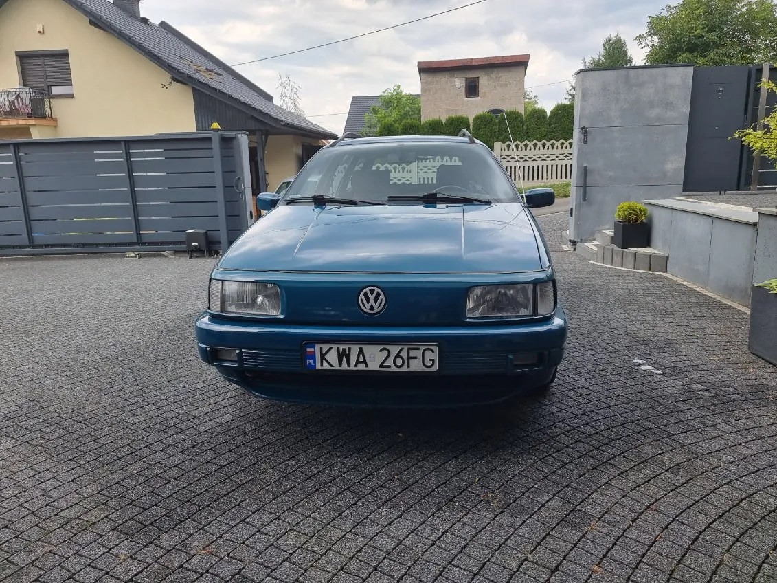 Niebieski samochód marki Volkswagen z przodu, z tablicą rejestracyjną KWA 26FG (z flagą PL po lewej stronie). Samochód stoi na brukowanej nawierzchni. W tle widoczne są domy: żółty z ciemnoszarym ogrodzeniem po lewej, szary budynek i biała palisada po prawej. Na niebie chmury, a na przedniej szybie samochodu widoczne są dekoracyjne naklejki. Wokół rosną krzewy i drzewa.