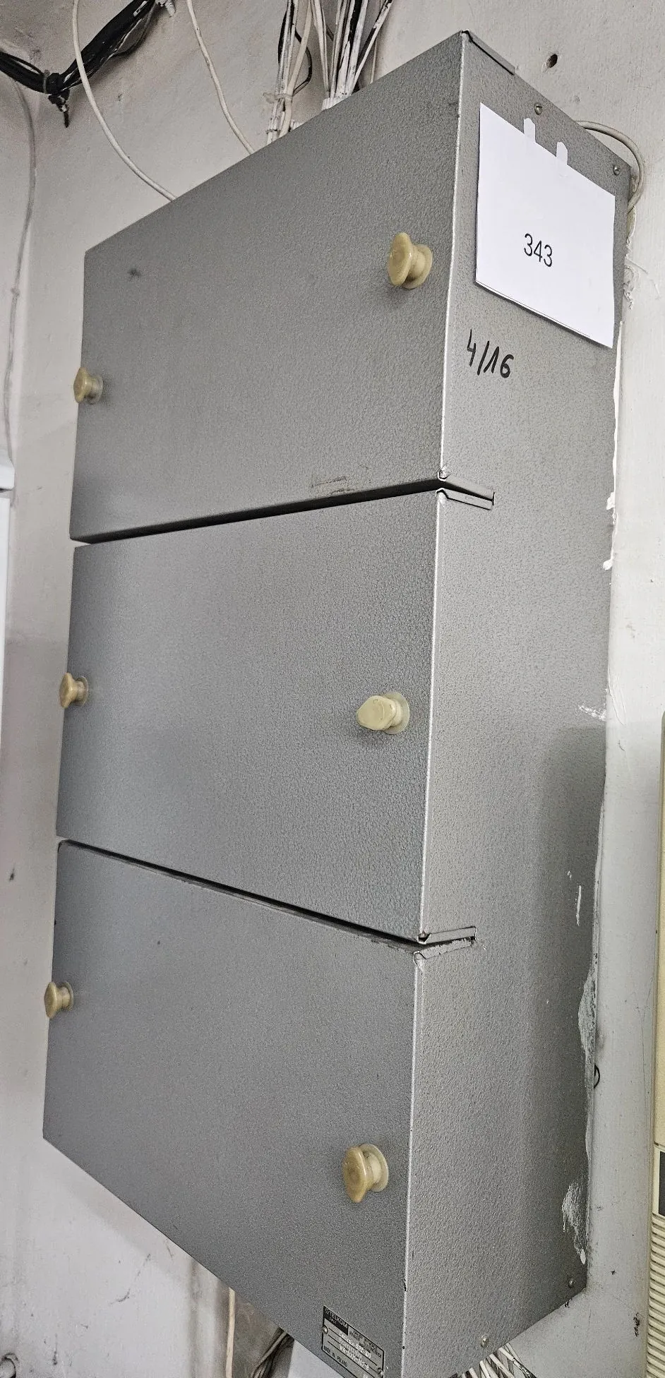 Srebrna, metalowa szafka z trzema szufladami zamontowana na białej ścianie. Każda szuflada posiada dwa małe, jasne uchwyty. Na górnej szufladzie przyklejona jest biała kartka z liczbą '343', a obok niej widnieje odręczny napis '4/16'. Nad szafką widoczne są zwisające kable elektryczne.