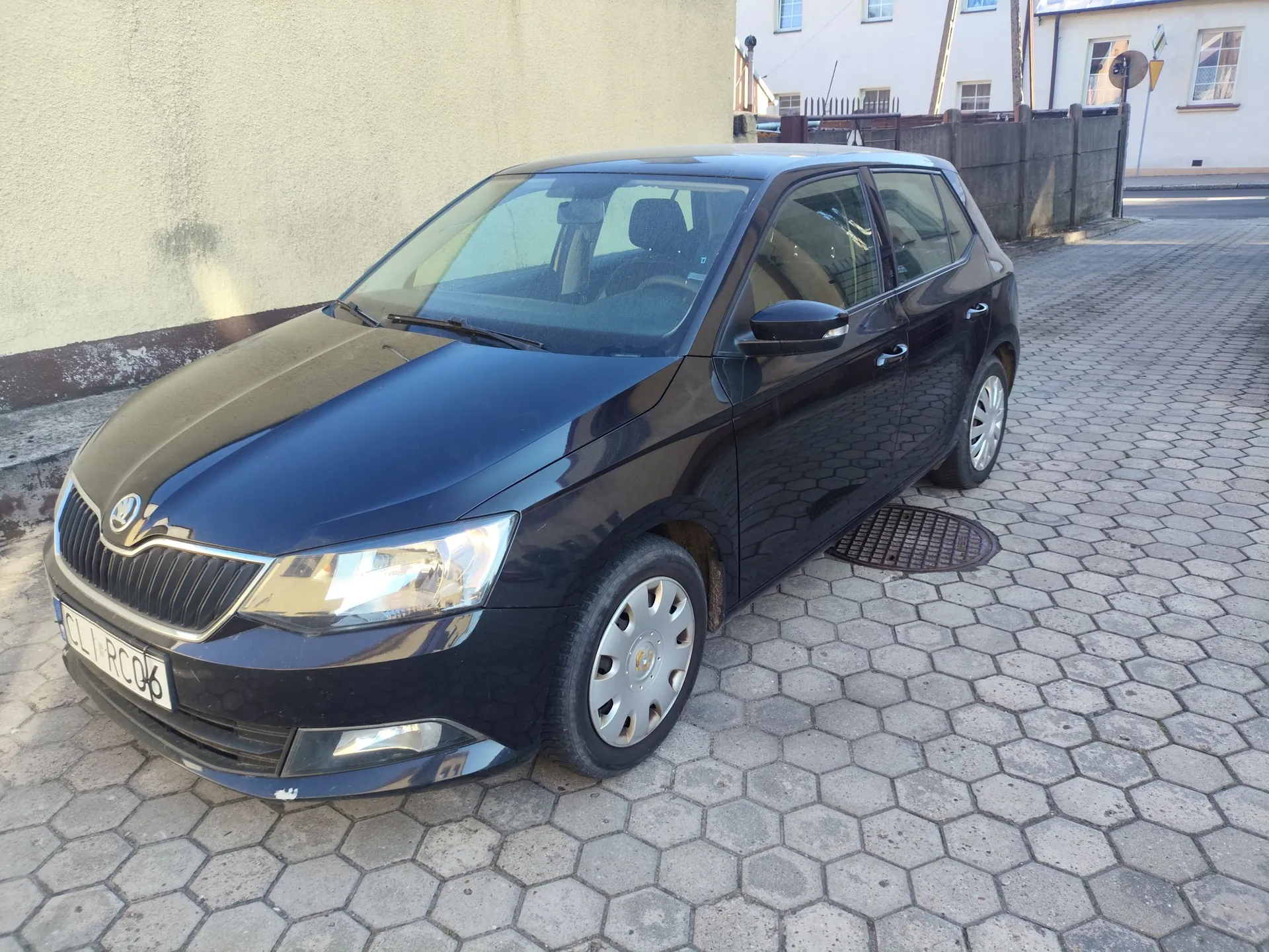 Czarny samochód marki Skoda zaparkowany na podjeździe z szarej, heksagonalnej kostki brukowej. Auto jest ustawione pod kątem, widoczny jest przód i prawy bok pojazdu. W tle znajduje się jasna ściana budynku oraz fragment ogrodzenia i innego budynku w oddali. Tablica rejestracyjna samochodu zawiera tekst: CLI RC06.