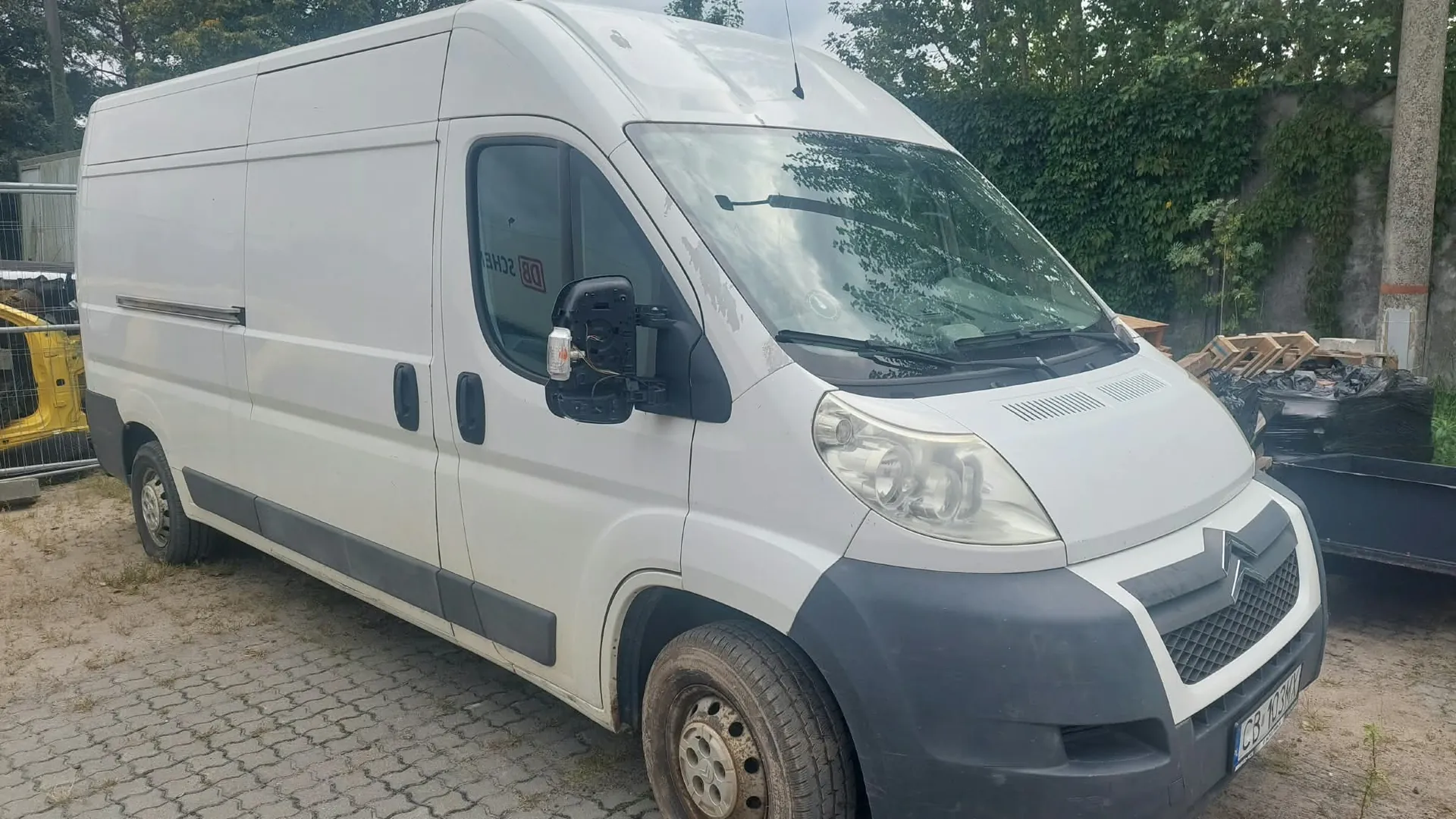 Biały furgon marki Citroën zaparkowany na kostce brukowej. Pojazd jest widoczny z przodu i z boku. W tle widać zielone krzewy, betonowy mur oraz fragmenty materiałów budowlanych i żółtej maszyny.