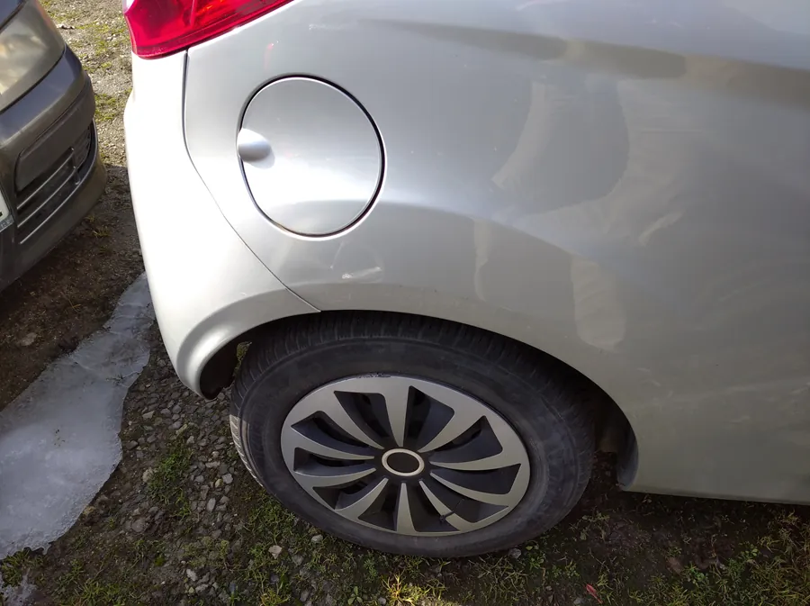 Zbliżenie na tylną część srebrnego samochodu, w tym koło z czarno-srebrnym felgiem o geometrycznym wzorze, okrągłą klapką zbiornika paliwa i czerwonym światłem tylnym. Po lewej stronie widoczna jest część przedniej osi innego, ciemnego samochodu. Podwozie stoi na podłożu z drobnych kamieni, trawą i fragmentami lodu.