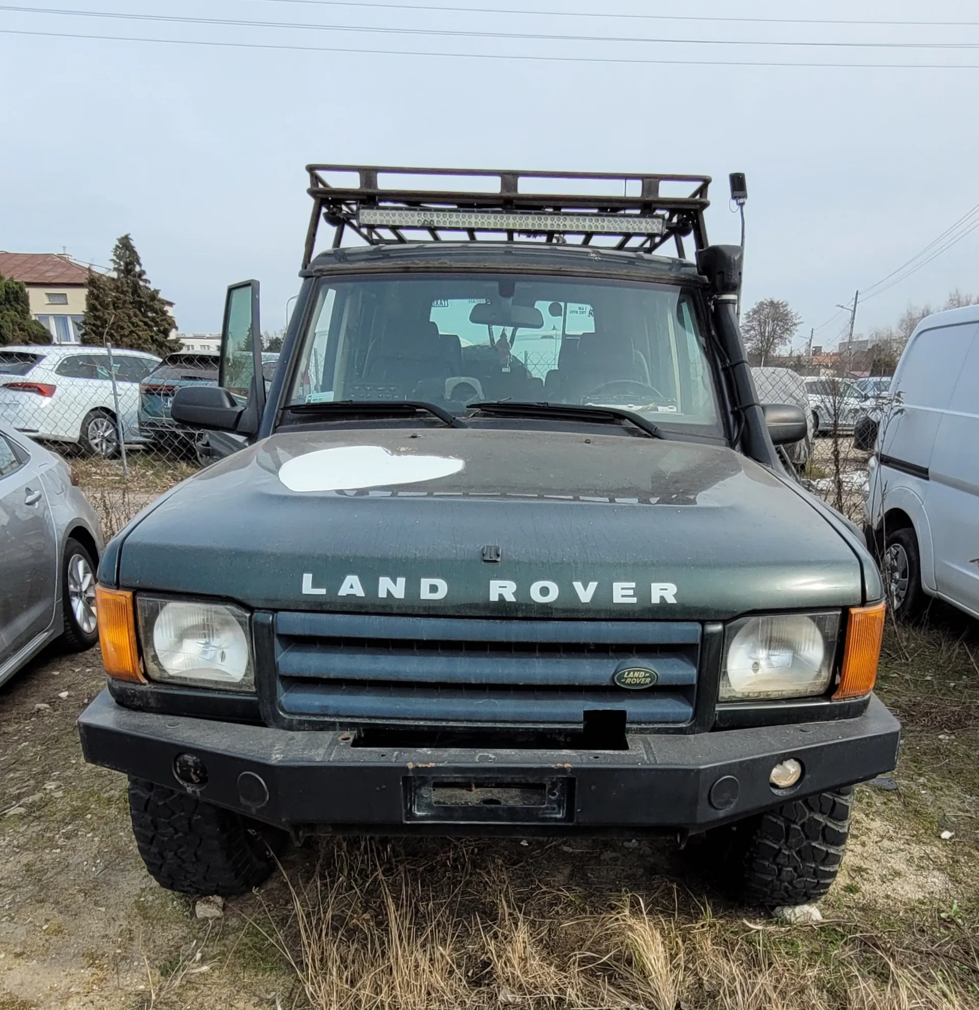 Przedni widok ciemnozielonego samochodu Land Rover z napisem "LAND ROVER" na masce. Pojazd wyposażony w dachowy uchwyt z listwą świetlną, otwarte drzwi kierowcy i czarny zderzak. Samochód stoi na trawiastym i ziemnym terenie, w tle widoczne są inne pojazdy (srebrny sedan, biały van), ogrodzenie siatkowe, budynki i drzewa. Niebo jest pochmurne. Na masce znajduje się biała plama, a tablica rejestracyjna jest pusta.