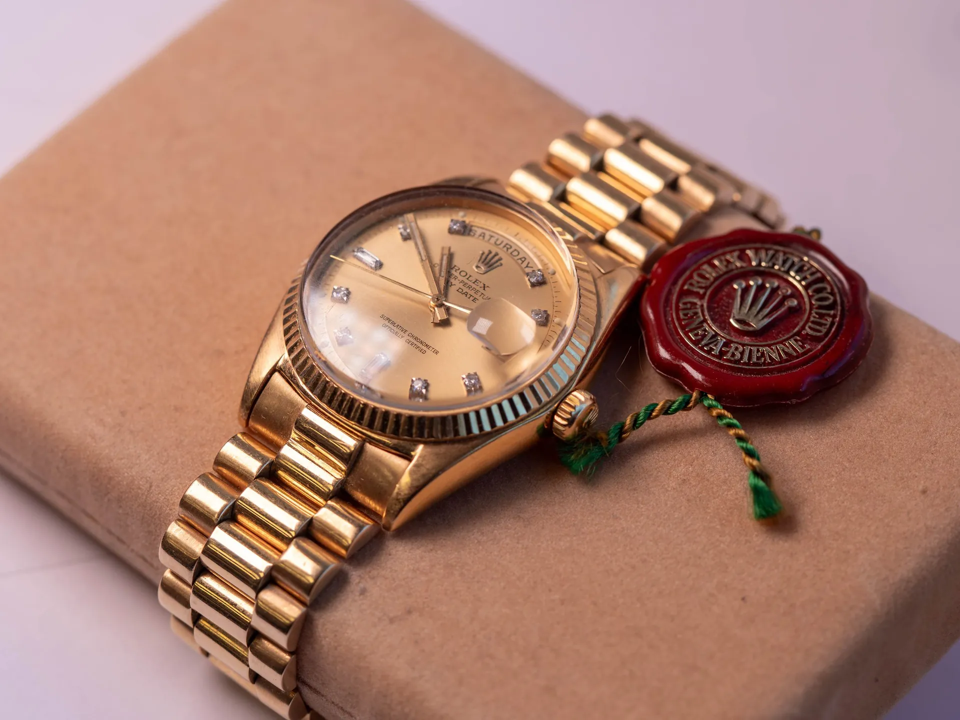 Złoty zegarek marki Rolex Datejust z metalową bransoletą, leżący na brązowej paczce zapakowanej w papier. Zegarek ma złotą tarczę z diamentowymi indeksami i datownikiem z lupą. Obok zegarka znajduje się czerwona lakowa pieczęć z logo korony i napisem „ROLEX WATCH CO. LTD GENEVE”, przymocowana zielono-żółtą sznurkiem.